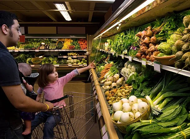 A family shopping in a grocery store produce section, with a man holding a baby and a child pointing to fresh vegetables, including onions, carrots, and greens.