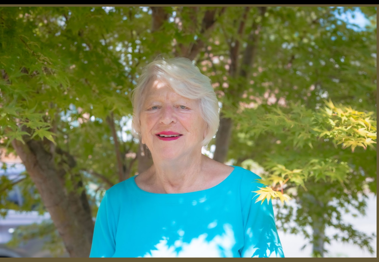 Elderly woman in a blue shirt standing outdoors with green leaves and trees in the background.