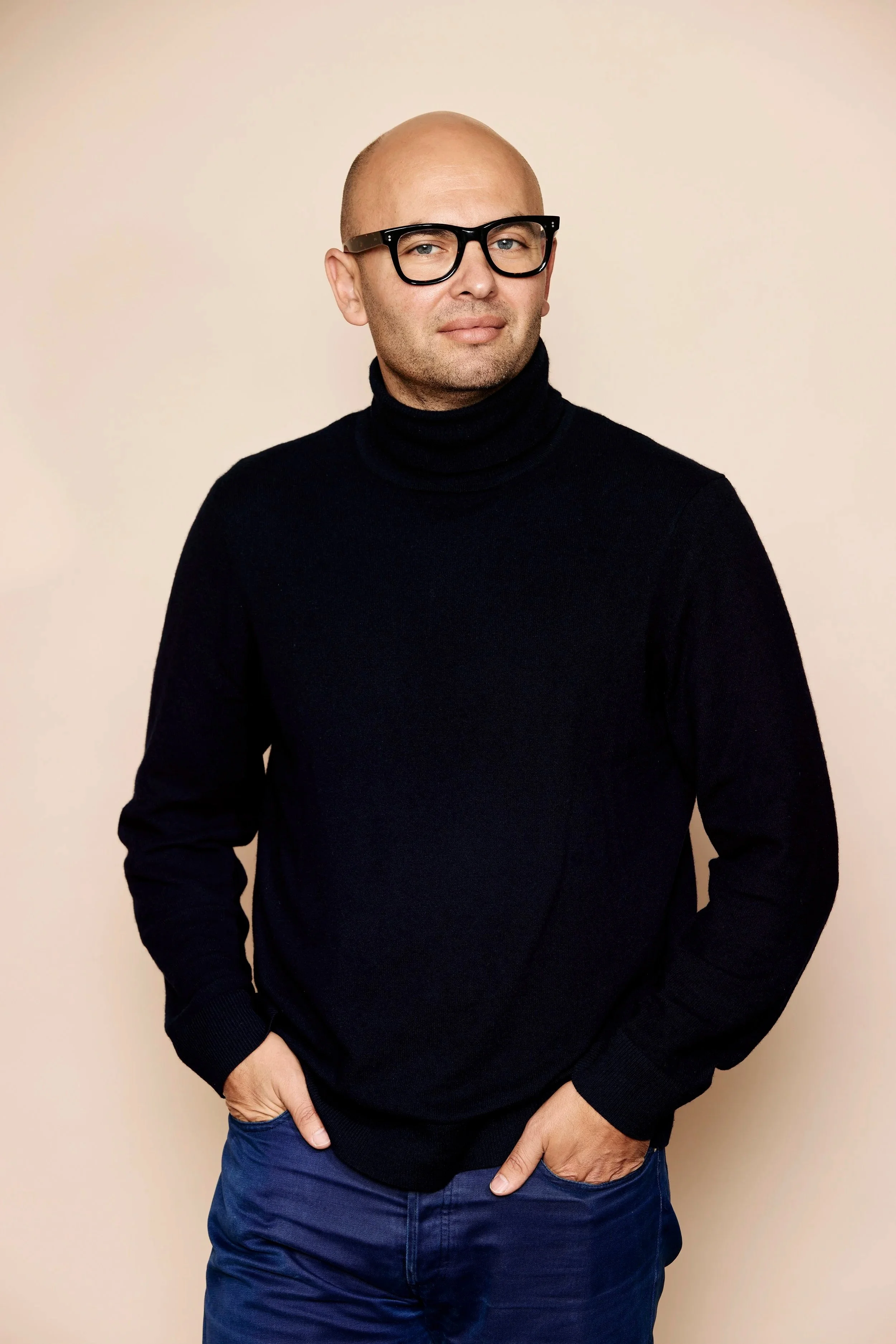 A man with a bald head, wearing black glasses and a black turtleneck sweater, standing against a beige background with his hands in his pockets.
