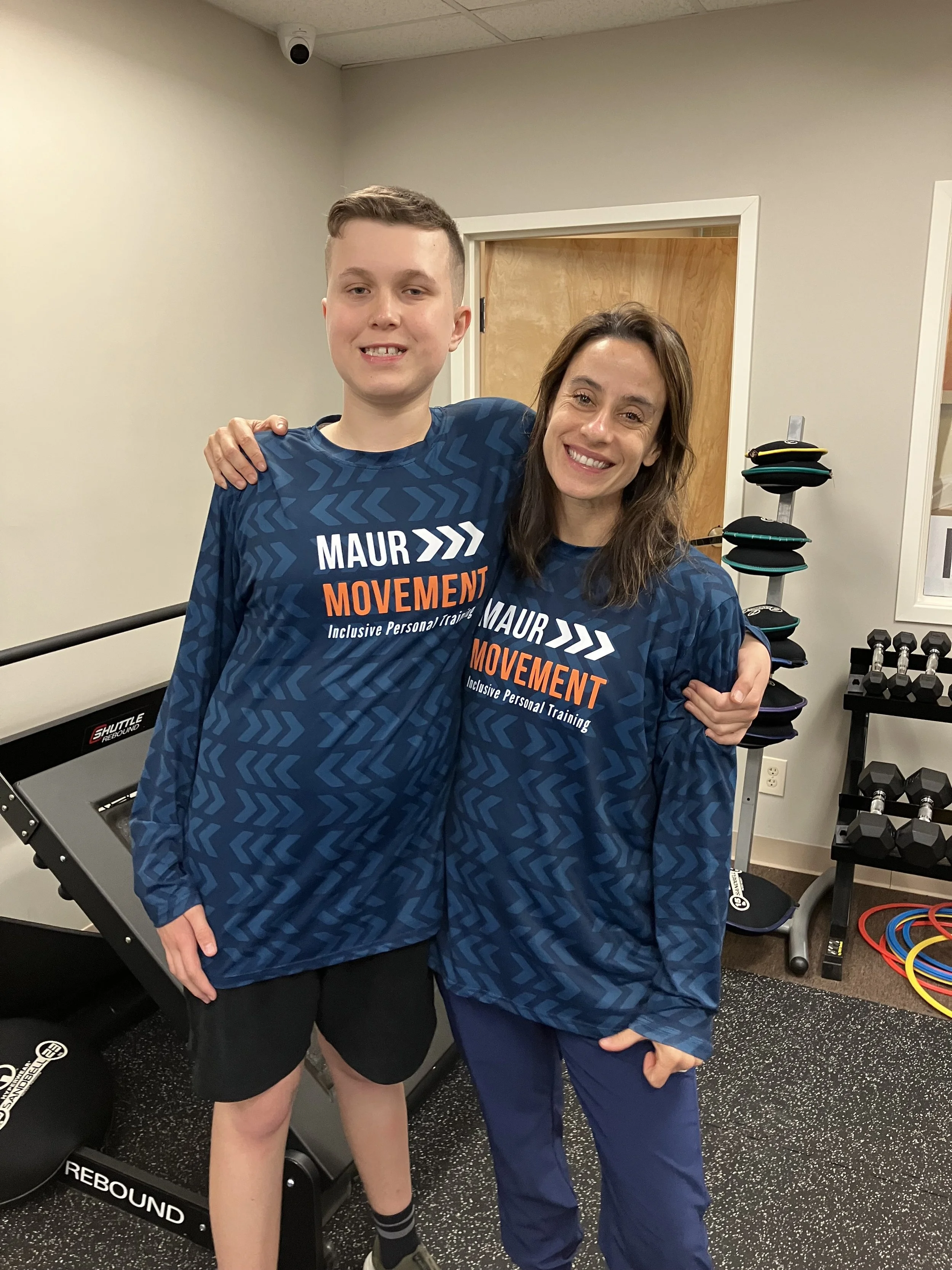 A young man and a woman smiling, standing together in a gym, both wearing matching blue workout shirts with 'MAUR MOVEMENT' printed on them.