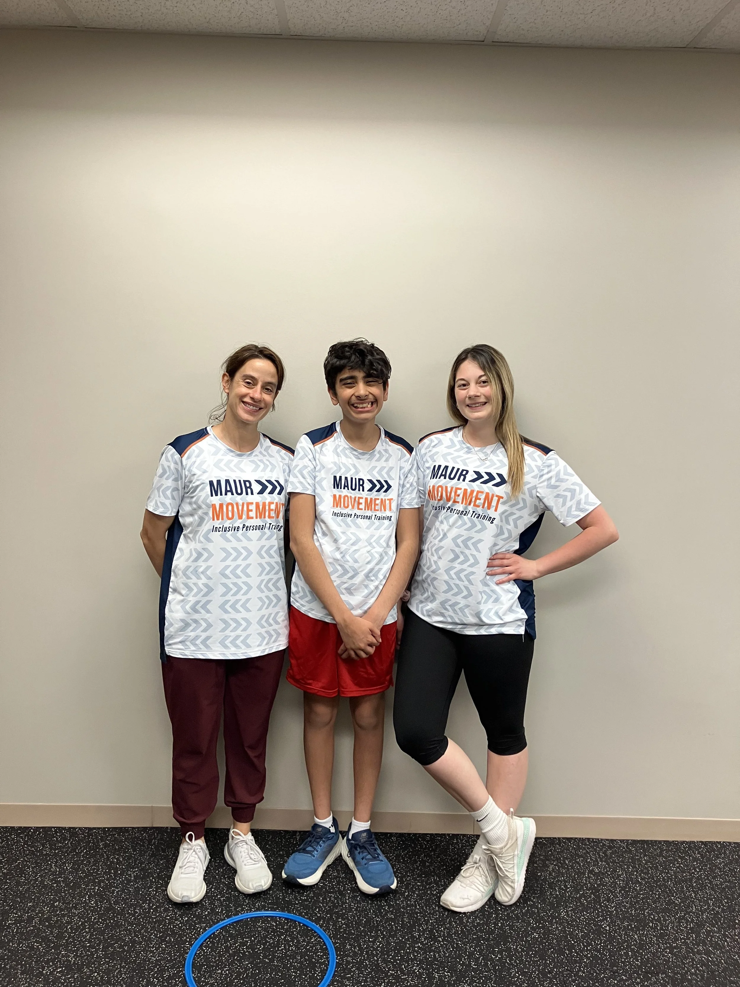 Three young women standing against a light-colored wall, wearing matching white athletic shirts with 'MAUR... Movement' printed on the front, smiling at the camera.