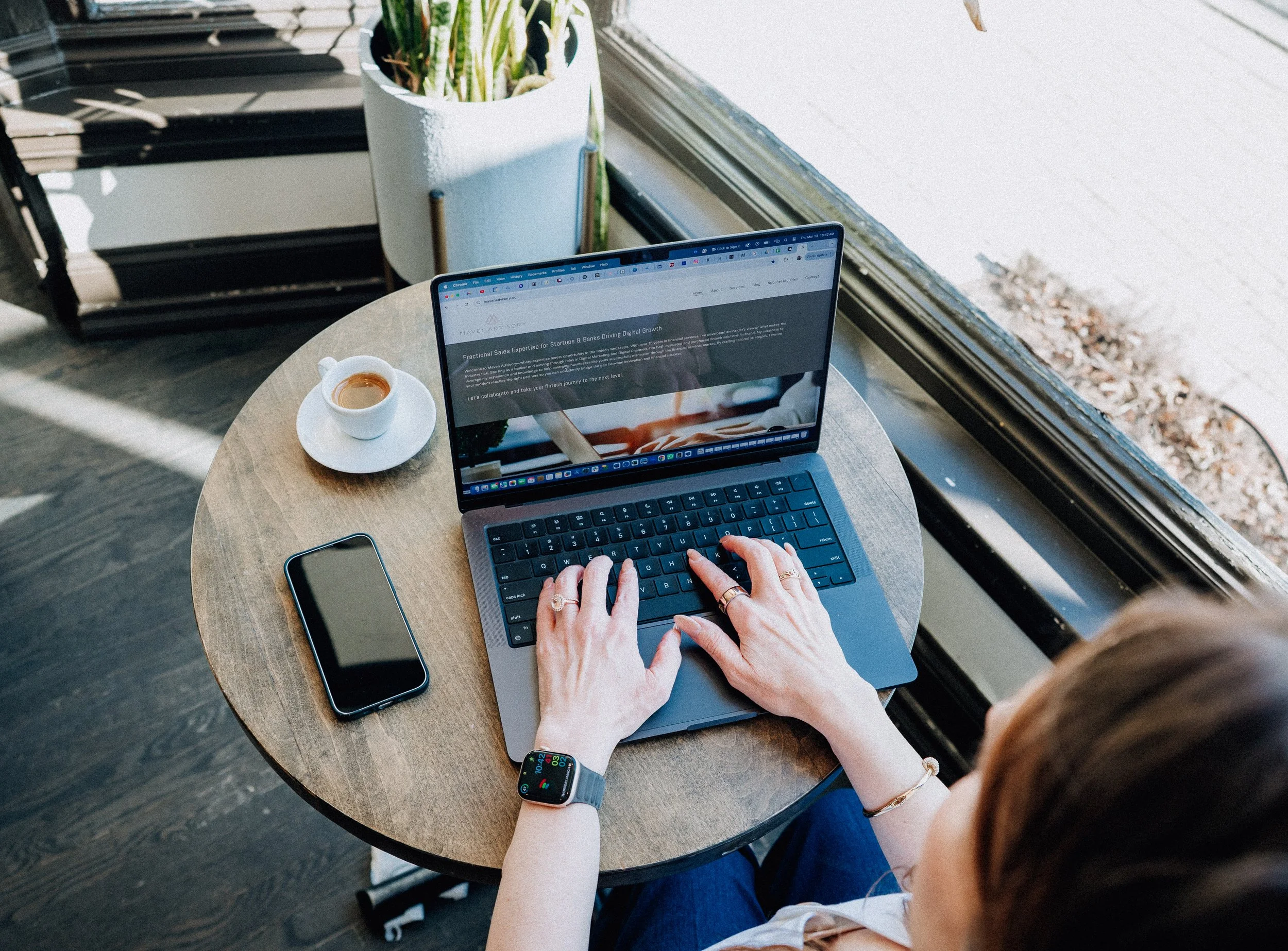 Angi Milano, Founder & CEO of Maven Advisory  with long brown hair using a laptop at a round wooden table by a window, with a coffee cup, a smartphone, a smartwatch, and a potted plant nearby.