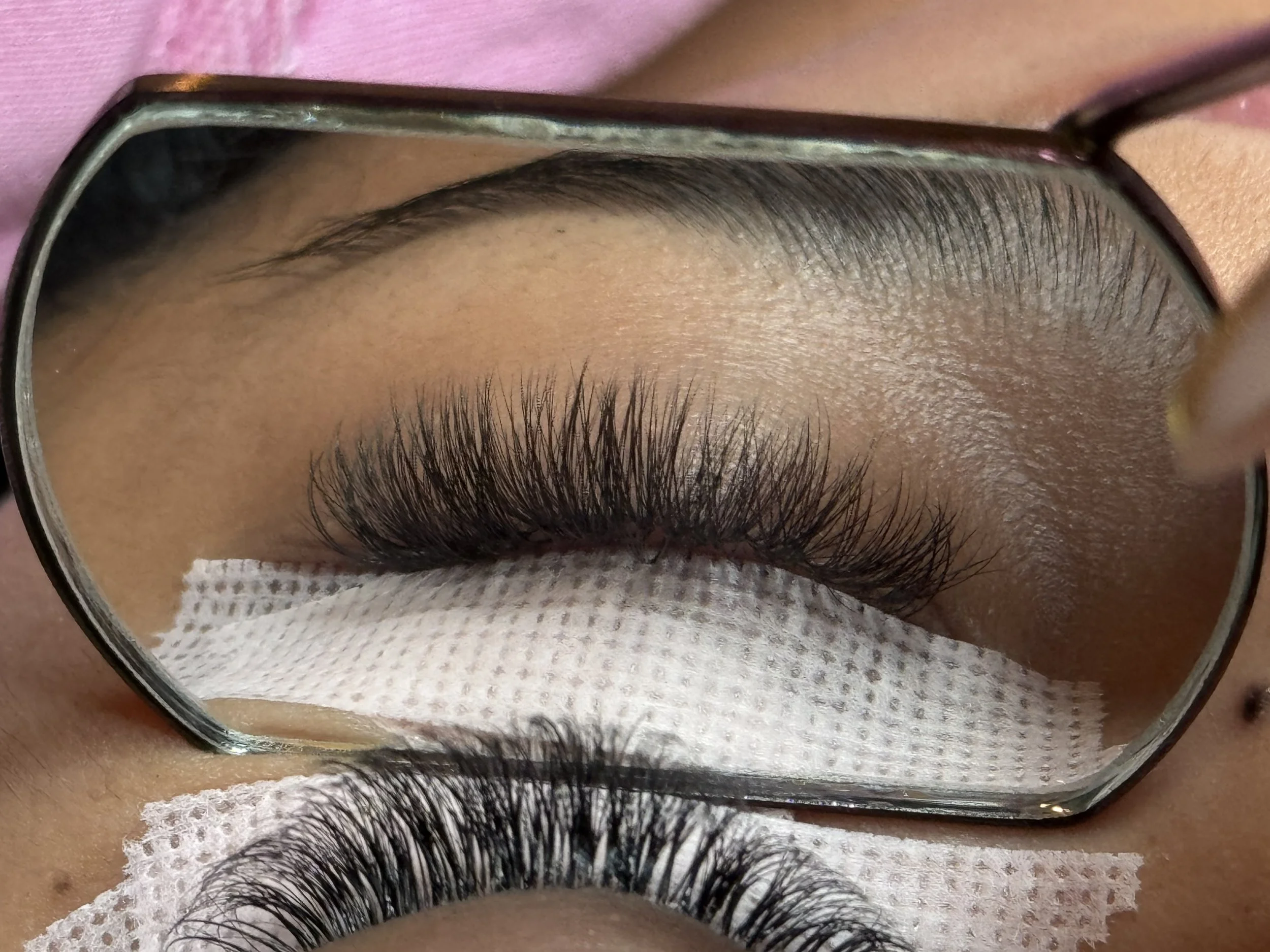 Close-up of a person's eye with long, curled eyelashes, seen through a magnifying glass. The person is wearing a pink cloth and a piece of white gauze under the eyelashes, possibly at a beauty or eyelash extension procedure.