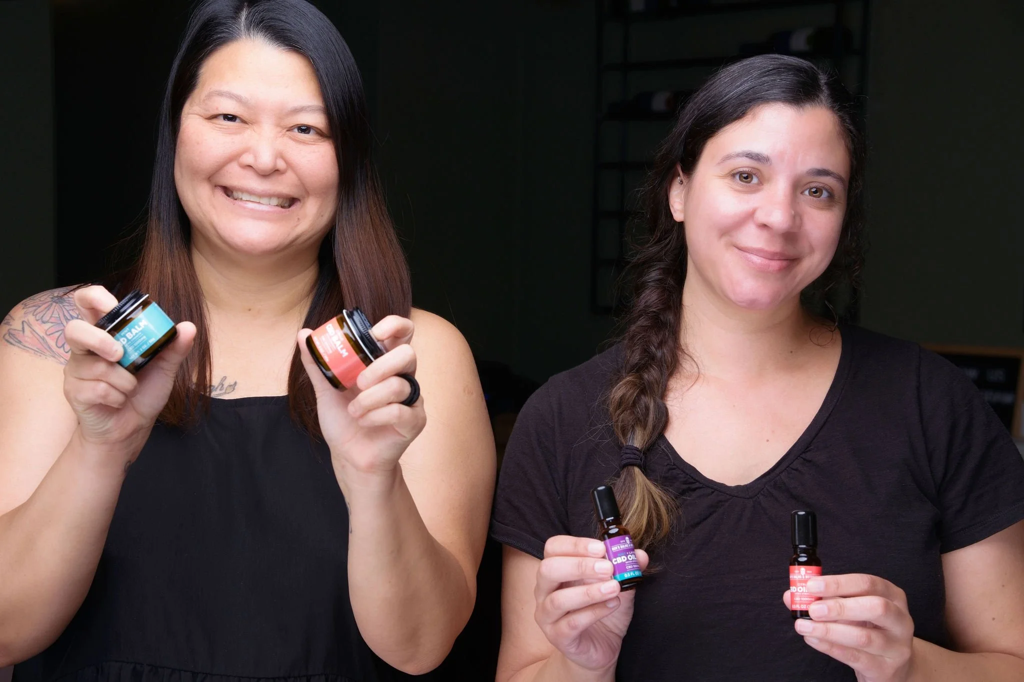 Two women smiling at the camera, each holding small bottles of CBD products. The woman on the left has long dark hair, a tattoo on her right shoulder, and is wearing a black sleeveless top. The woman on the right has long dark hair in a braid and is wearing a black t-shirt.