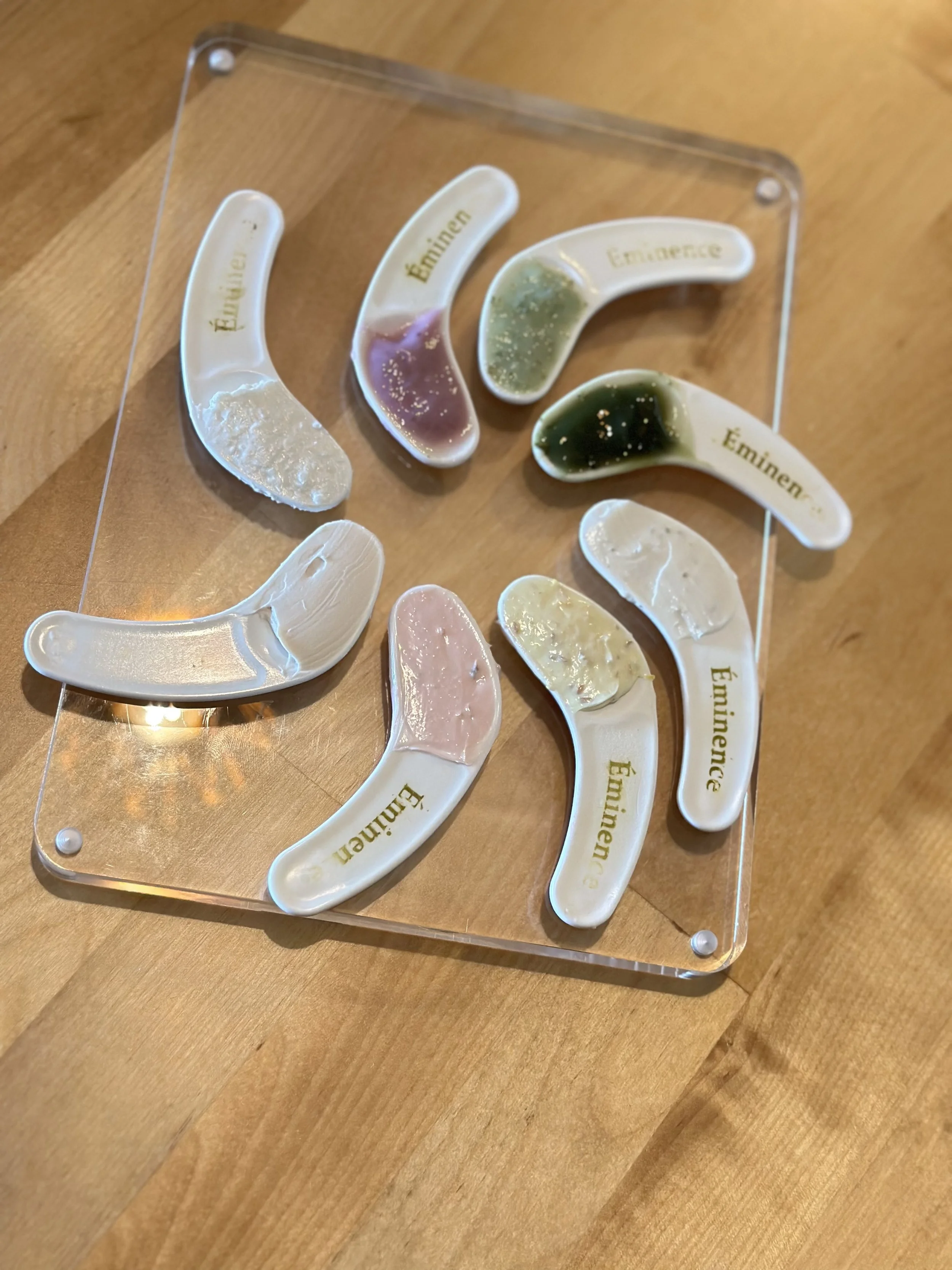 Display of six teardrop-shaped spoons with different colors of paint or sauce, each labeled with the names of rappers Eminem and Lil Wayne in gold writing, on a glass surface on a wooden table.