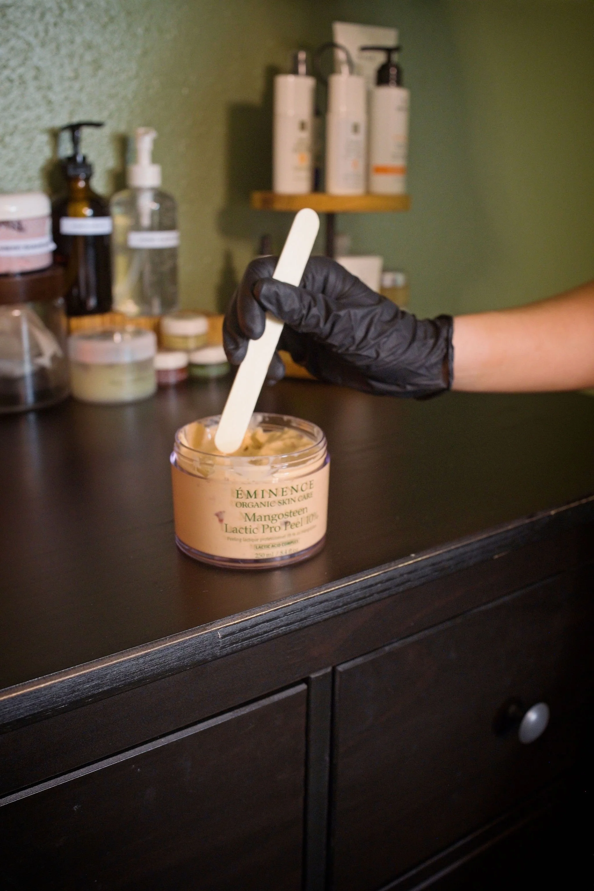 A person wearing black gloves stirring a skincare product in a jar labeled 'Eminence Organic Skin Care, Mangosteen Lactic Pro Peel 10%' using a wooden spatula, with various skincare bottles and jars in the background.