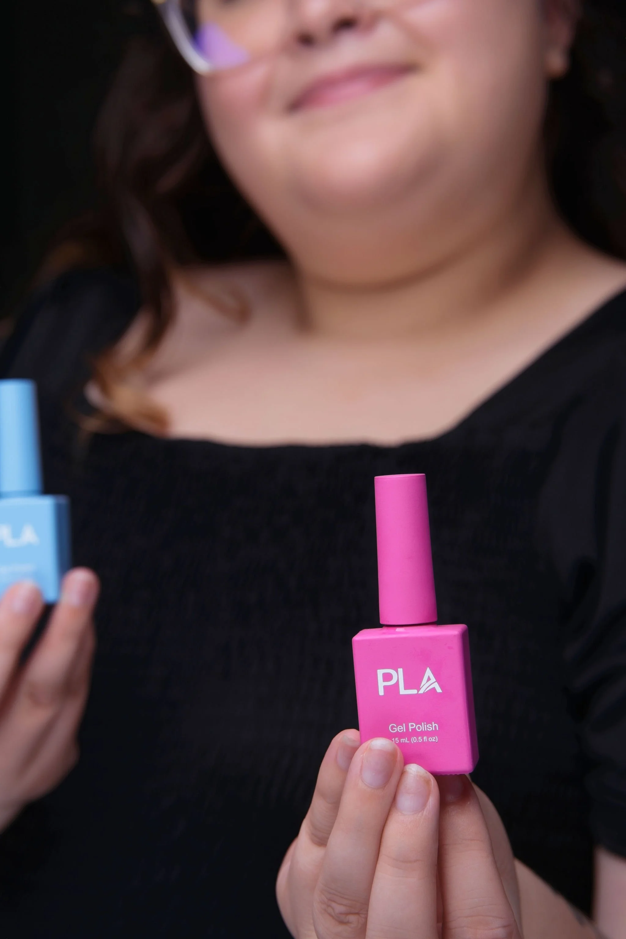 A woman holding a pink bottle of gel nail polish labeled 'PLA Gel Polish' with a woman in the background holding a blue bottle of nail polish.