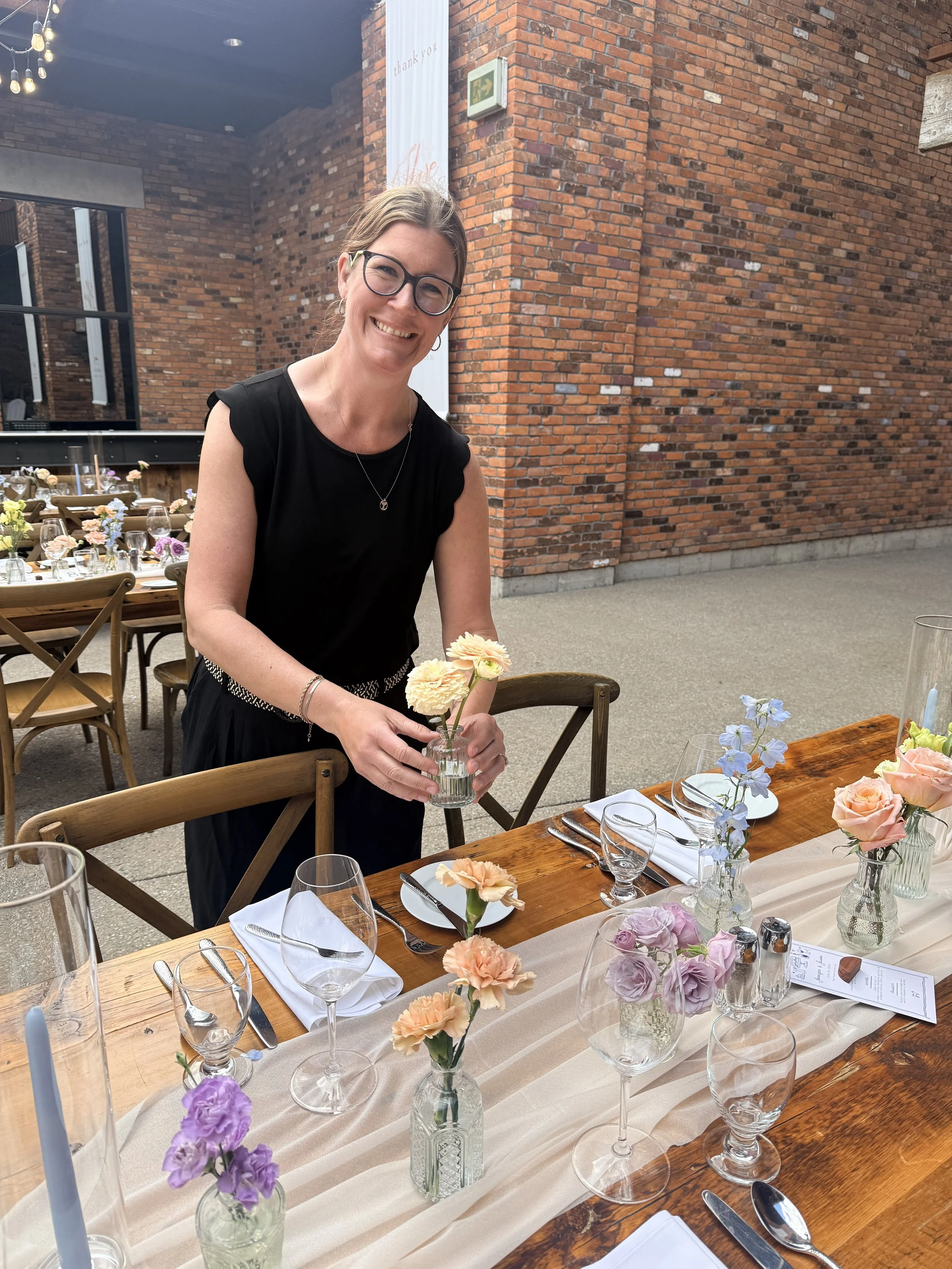 A smiling woman with glasses and earrings in a black dress holding a vase with cream-colored flowers at a decorated table set for a meal. The table has floral centerpieces, clear glassware, white napkins, and silverware. The setting is indoors with wooden tables, brick walls, and a large window or mirror behind her.