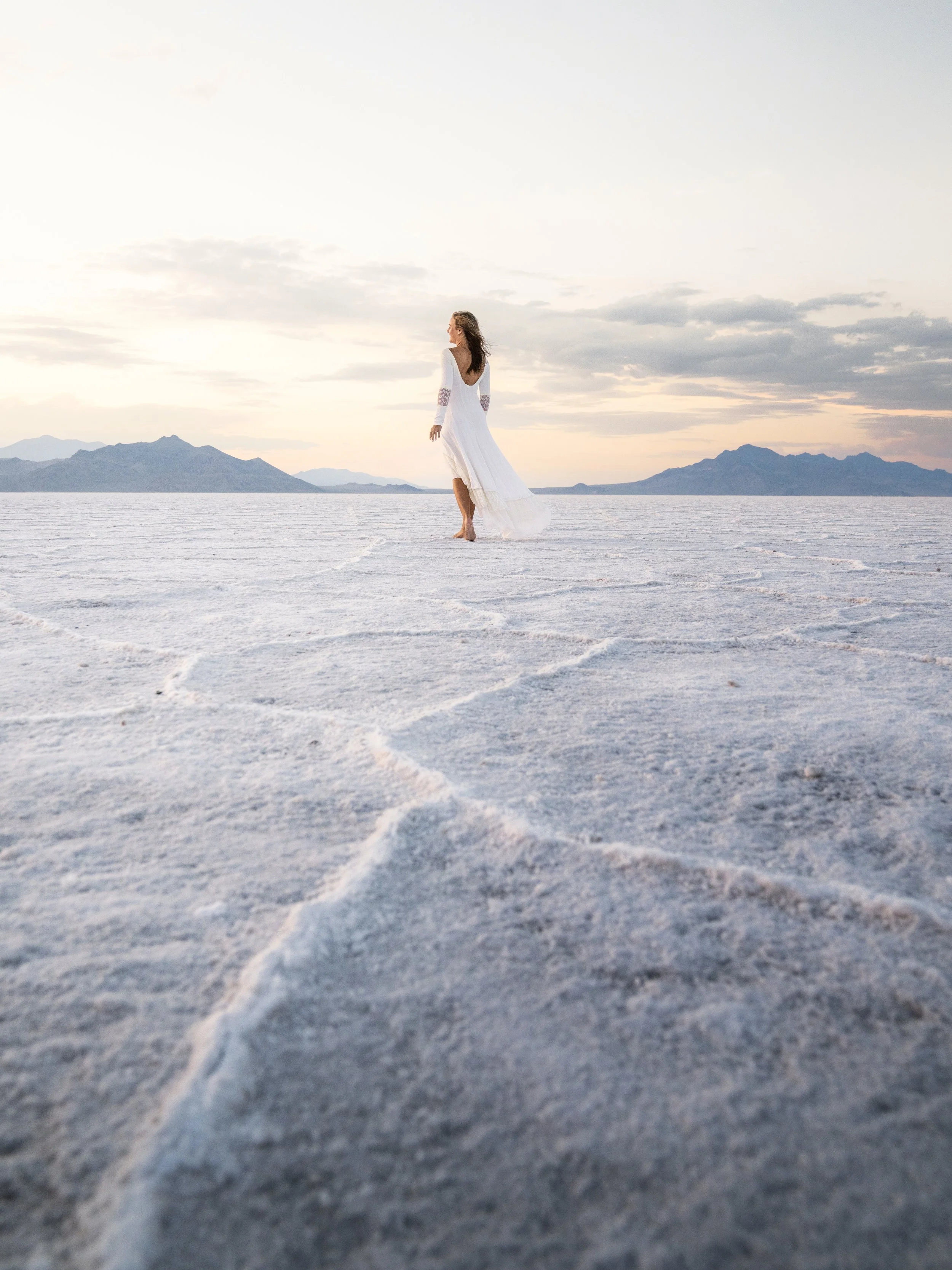 A woman in a white dress walking barefoot on a flat, salt flat terrain at sunset with mountains in the distance.