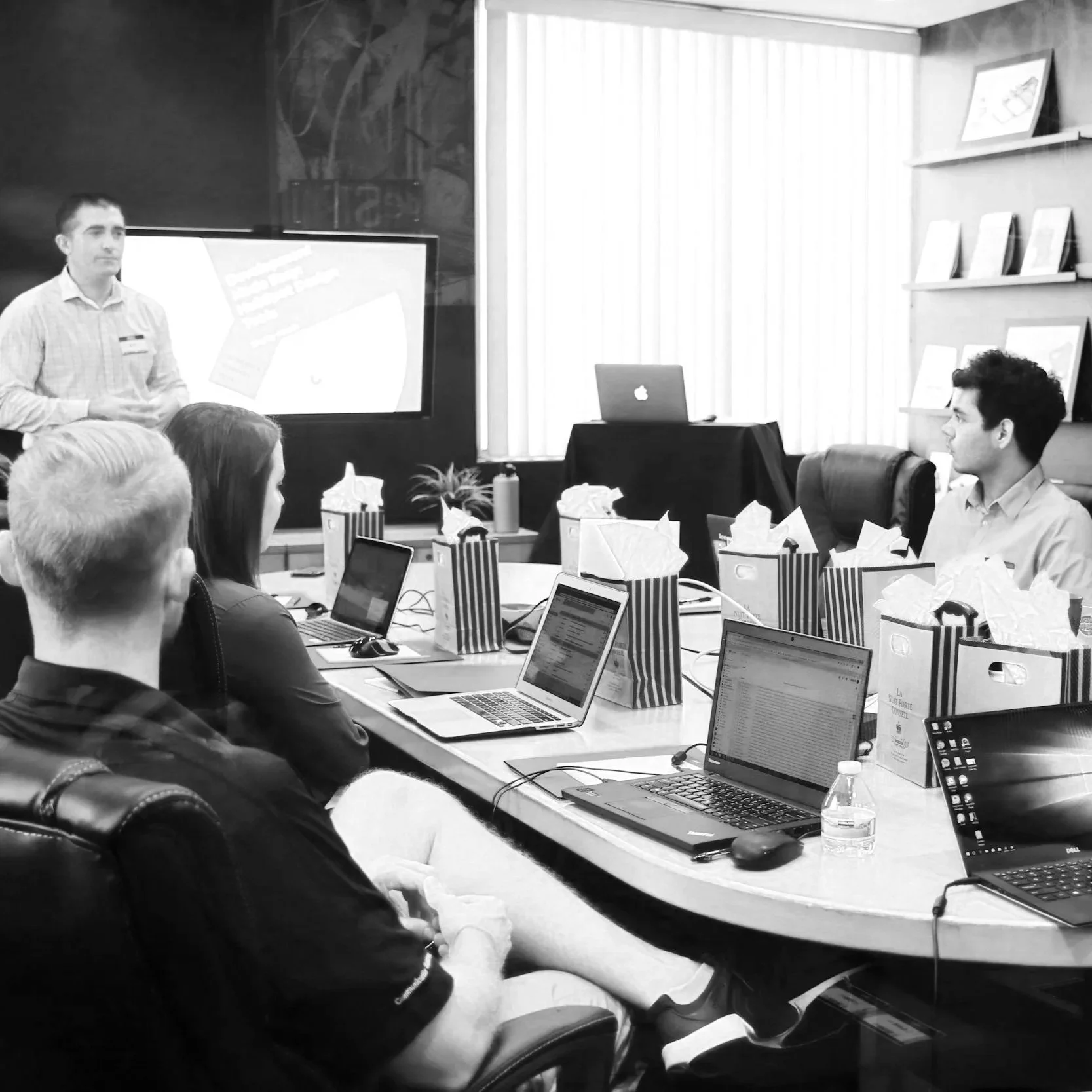 A group of people in a conference room, sitting around a long table with laptops, papers, and water bottles, while a presenter stands near a screen at the front of the room.