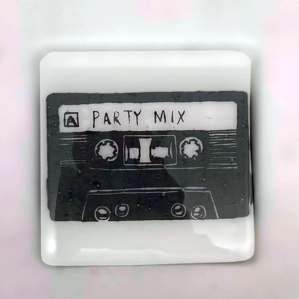 Overhead view of a white kiln-formed glass coaster with a black enamel retro cassette tape Party Mix design