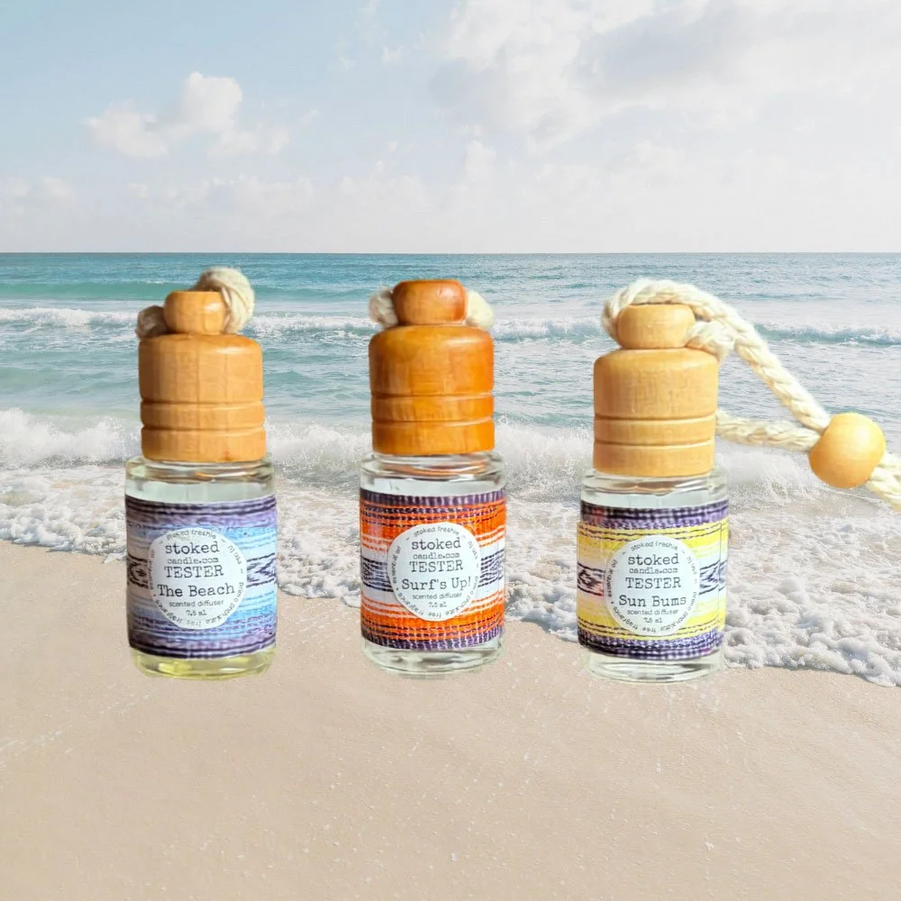 Three Stoked Freshie scented hanging diffusers labeled The Beach, Surf's Up, and Sun Bums standing in beach sand with ocean waves