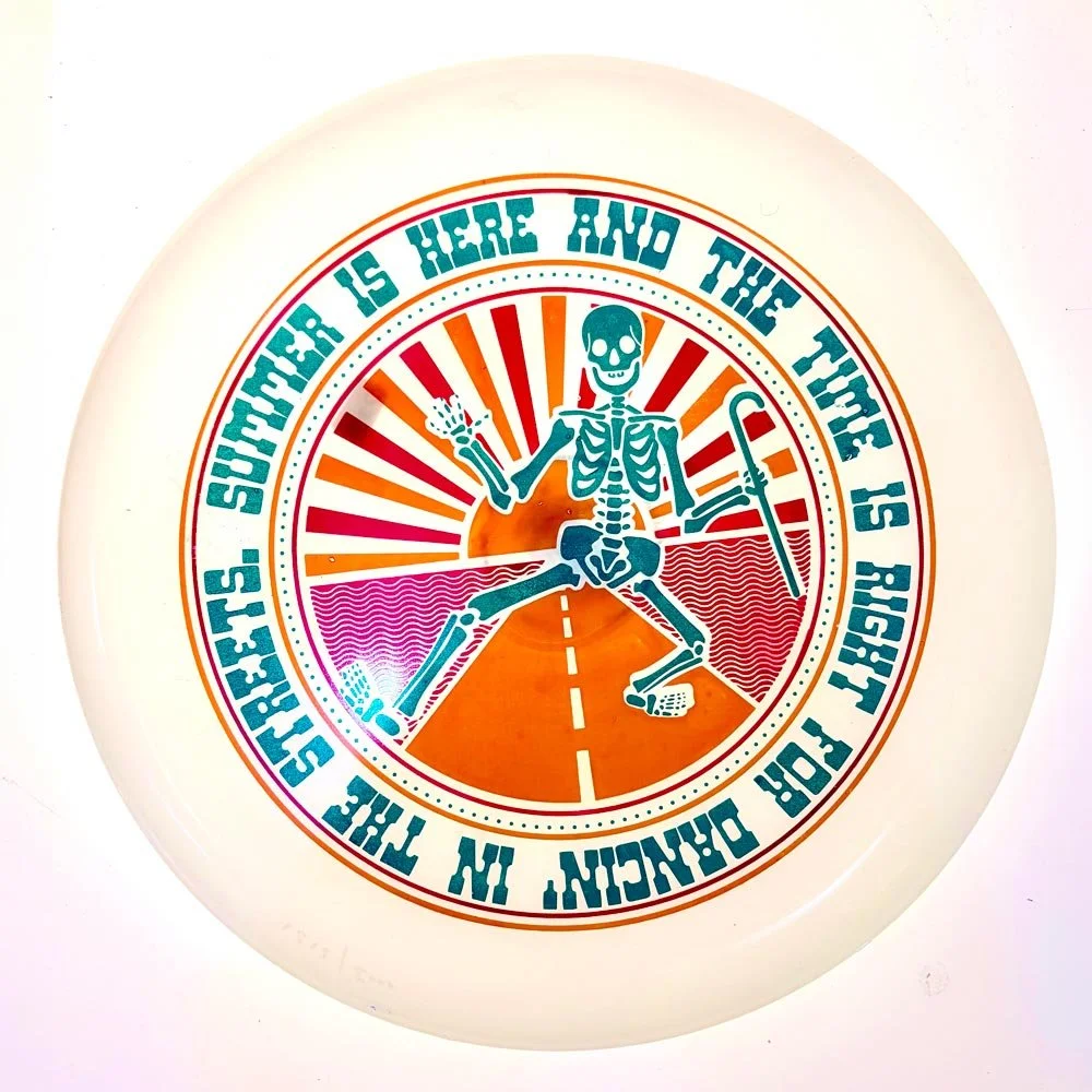 White glow frisbee with teal dancing skeleton on orange sunburst design with colorful lyric text border