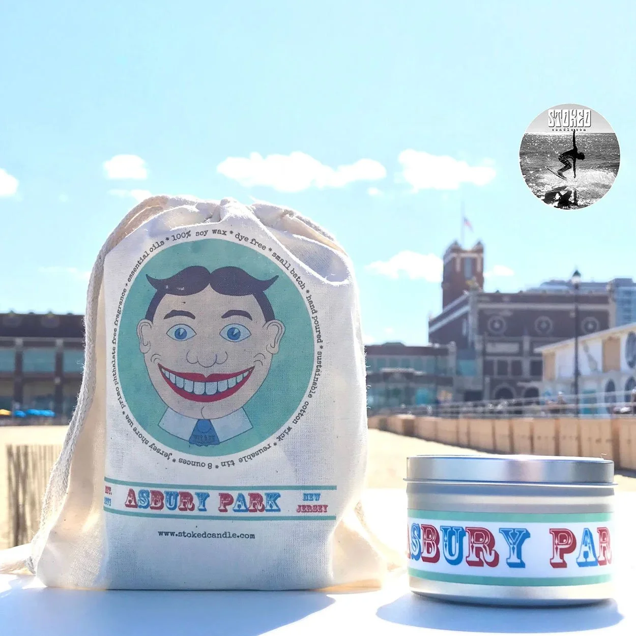 Stoked Candle Tillie Asbury Park tin and drawstring bag on boardwalk with Asbury Park Convention Hall in background