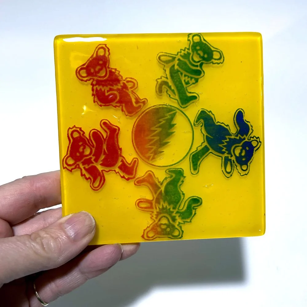 Handheld view of a yellow fused glass coaster with multicolored dancing bears circling a lightning bolt design