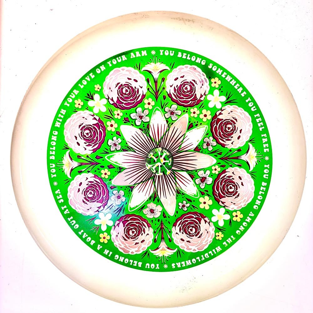 White glow frisbee with vibrant green floral mandala featuring roses and a passionflower with lyric text border