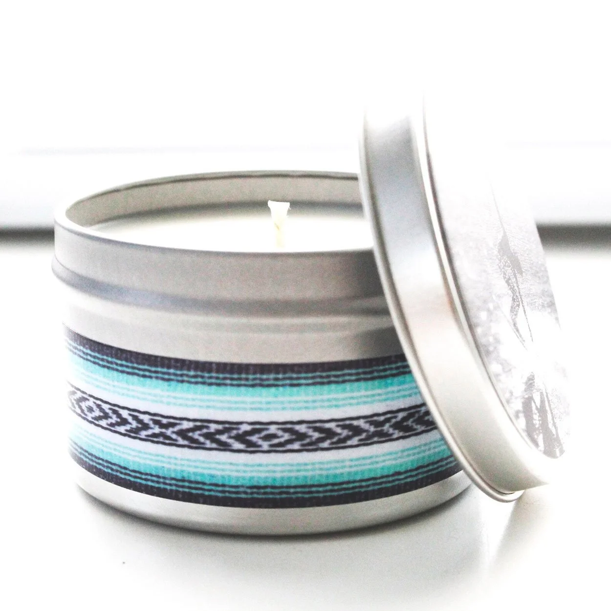 Stoked Candle The Beach tin with teal and black baja blanket print label and lid leaning against it on white background