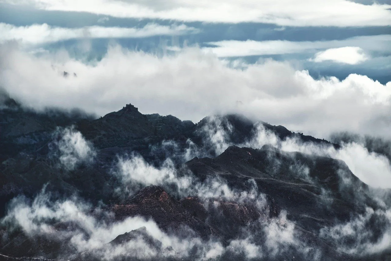 Mountain range with dark, rugged peaks partially obscured by swirling clouds and mist under a partly cloudy sky.