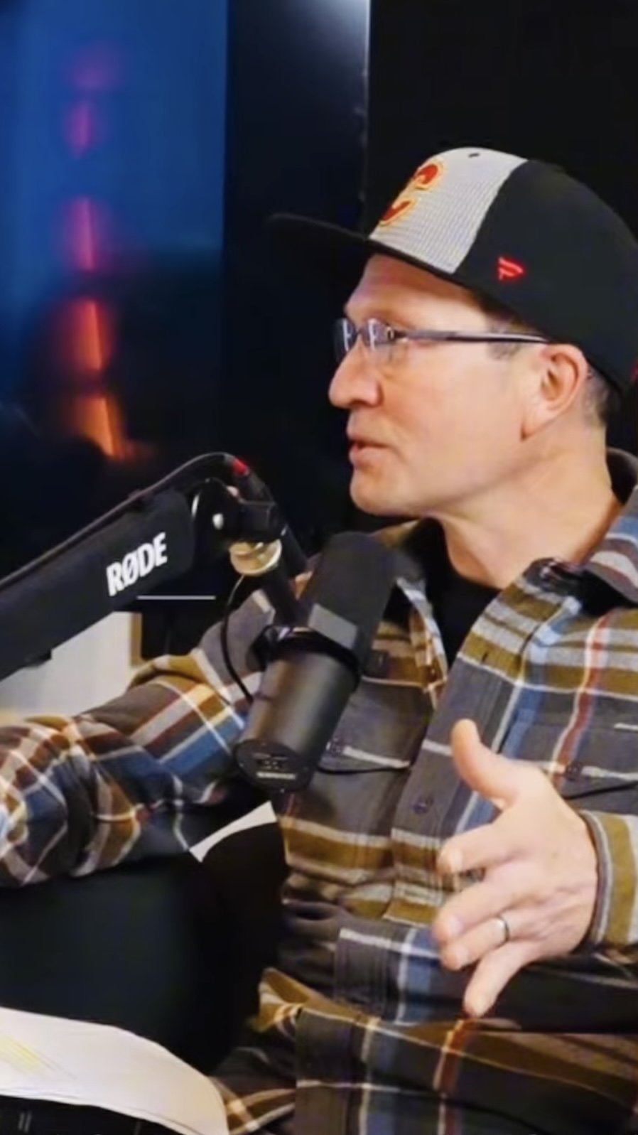 A man wearing glasses and a baseball cap with a graphic design, sitting at a table with a microphone labeled RODE, gesturing with his right hand, in a radio or recording studio.