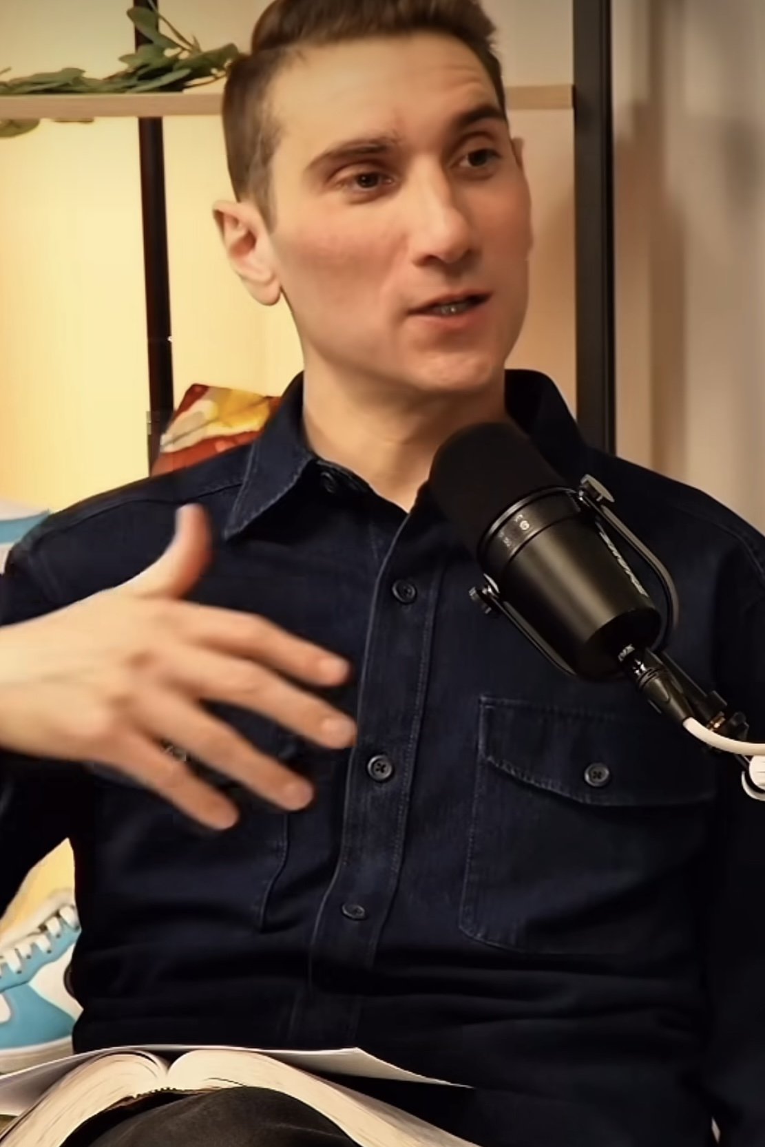 A young man with short brown hair and fair skin speaking into a microphone, wearing a dark denim shirt. He is gesturing with his hand on his chest, sitting indoors near a shelf with some plants behind him.