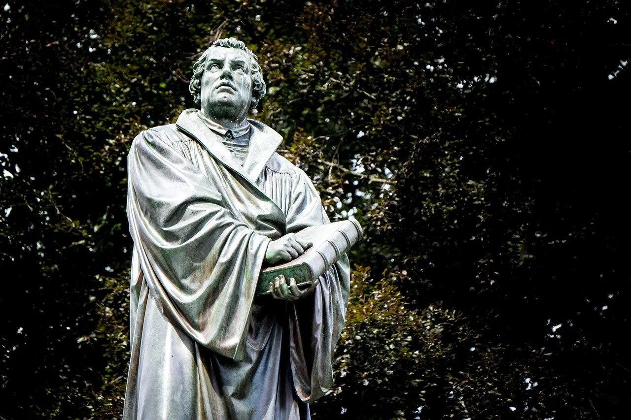 A statue of a historical figure wearing academic robes, holding a rolled-up document, with a serious expression, set against a background of dark green trees.