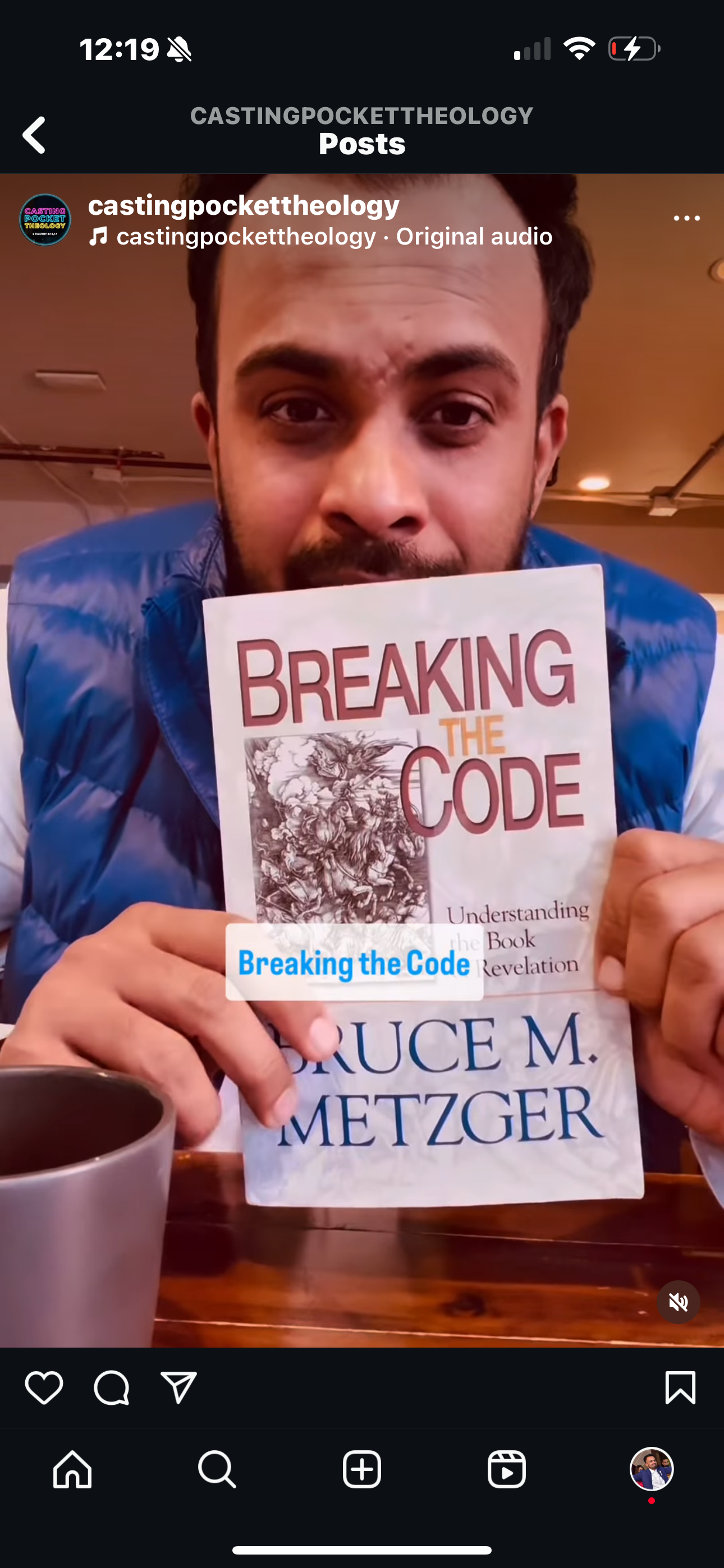 A man is holding a book titled 'Breaking the Code' by Bruce M. Metzger, partially covering his face. He is wearing a blue jacket and sitting at a table with a coffee mug in view.