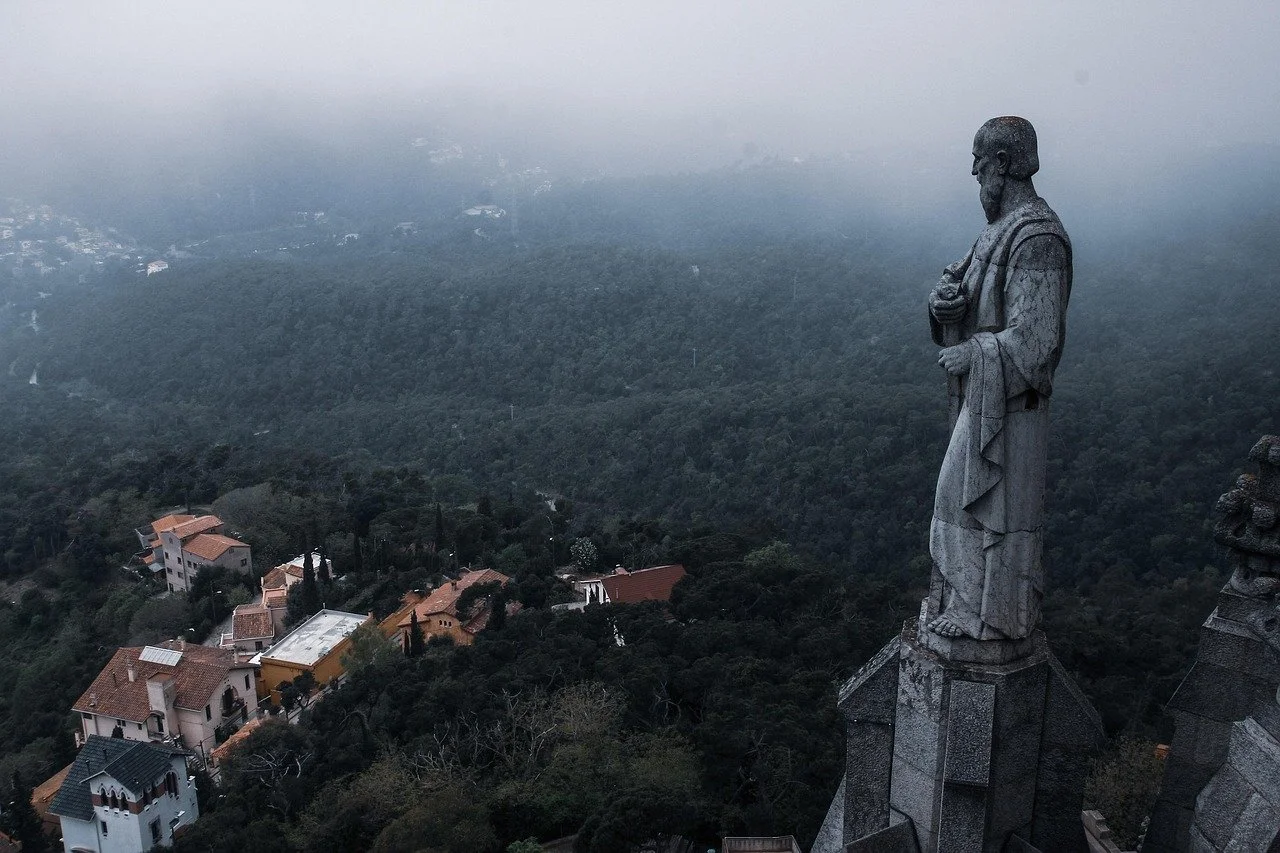 A large stone statue of a man stands overlooking a foggy landscape with trees and houses below.