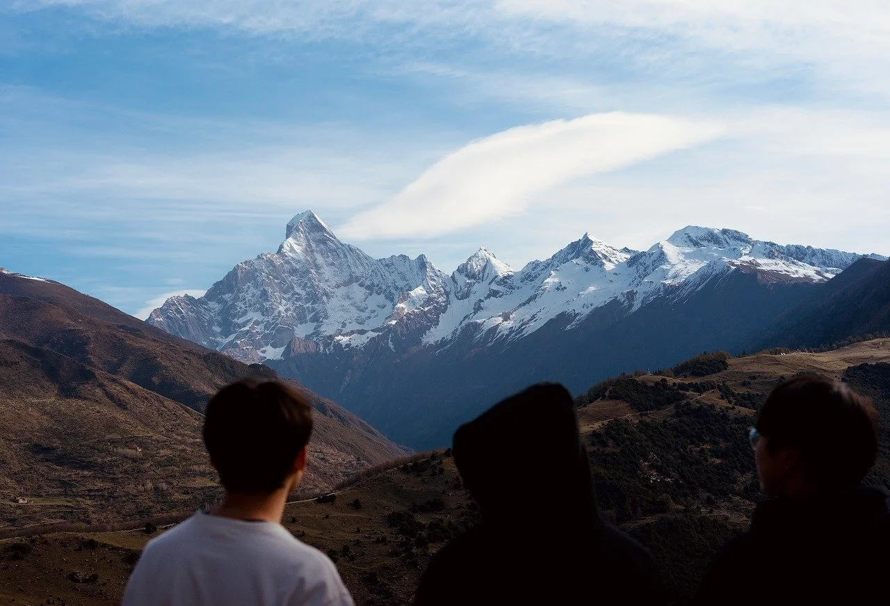 Three people looking at snow-capped mountains in the distance with a partly cloudy sky.
