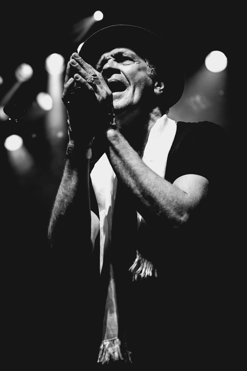 A black and white photo of a male singer passionately performing on stage, wearing a hat and a scarf, singing into a microphone with his eyes closed and hands clasped.