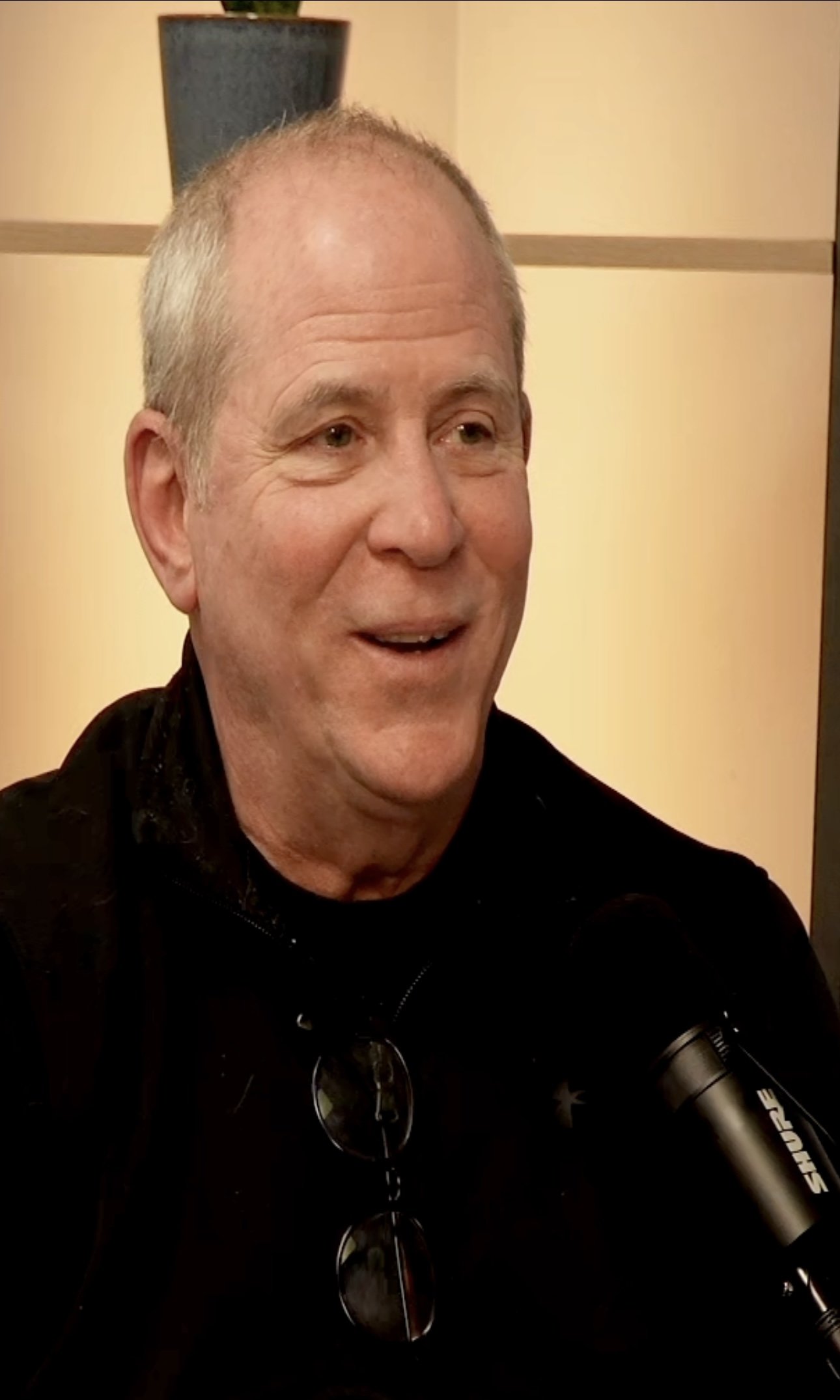 A middle-aged man with short, graying hair smiling while speaking into a microphone. He is wearing glasses hanging from his neck and a black jacket. There is a navy blue pot with a green plant on a shelf behind him and beige-colored walls.