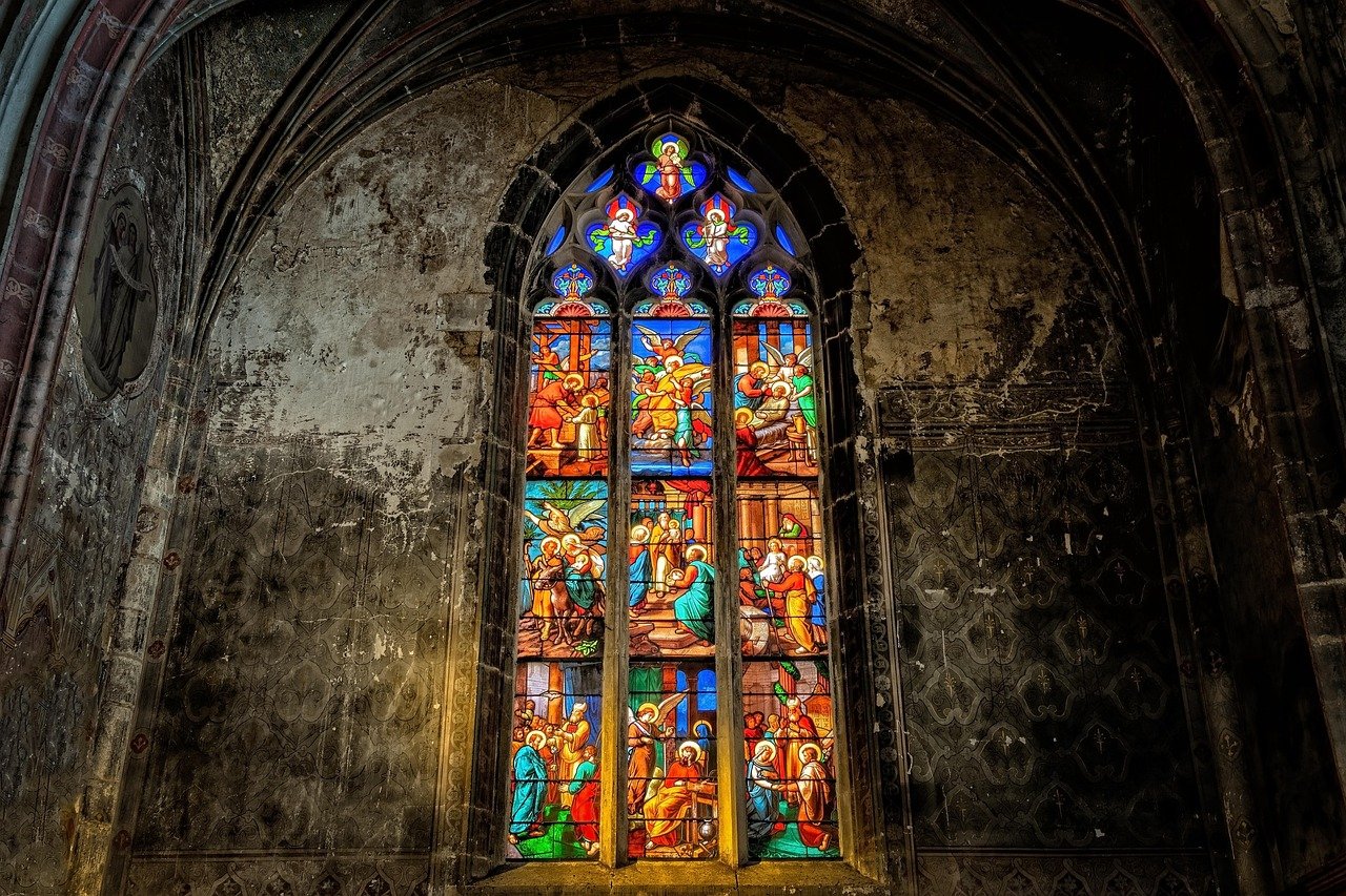 Colorful stained glass window with biblical scenes, set in a dark, weathered stone wall of a church.