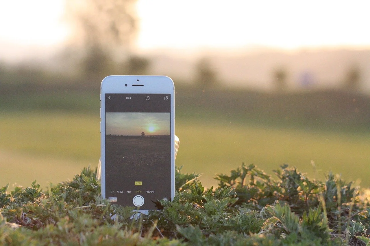 Smartphone standing upright on grass, capturing a photo of a sunset over a rural landscape.