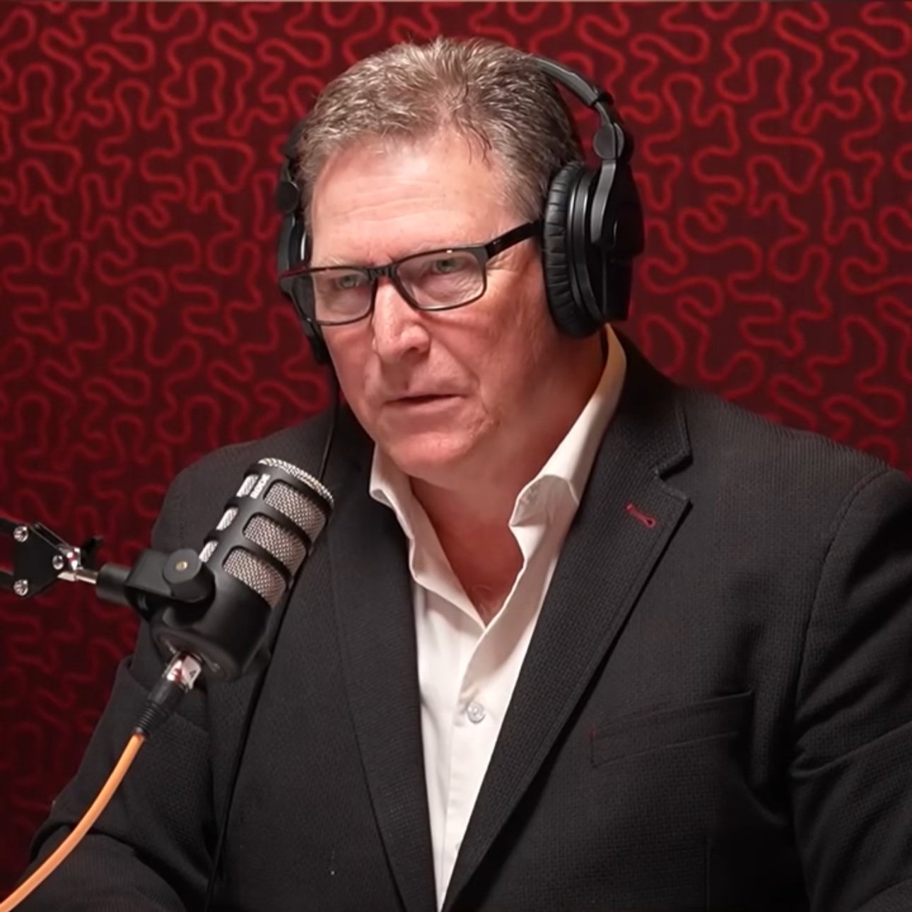 A man with gray hair, glasses, and a suit jacket, wearing headphones, speaking into a microphone in a recording studio with red soundproof wall panels.