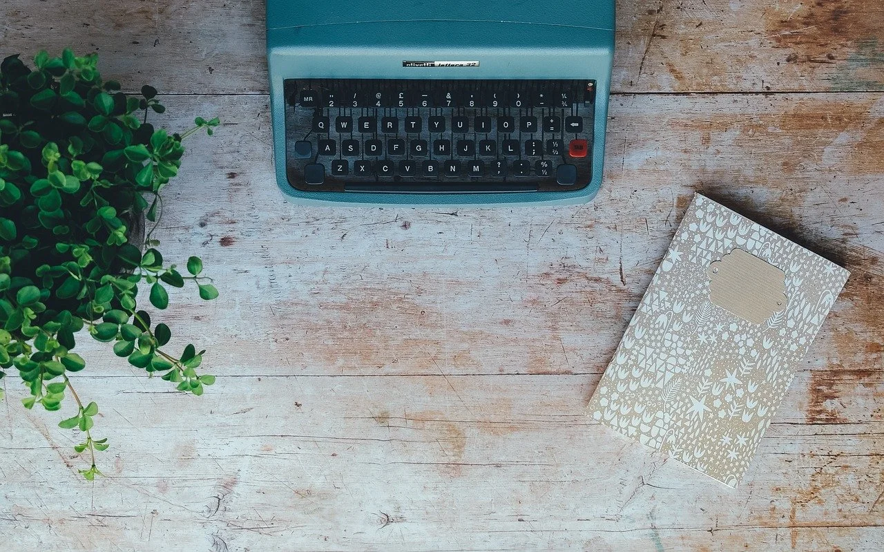 A vintage typewriter, a notebook with decorative patterns, and a potted plant on a rustic wooden desk.