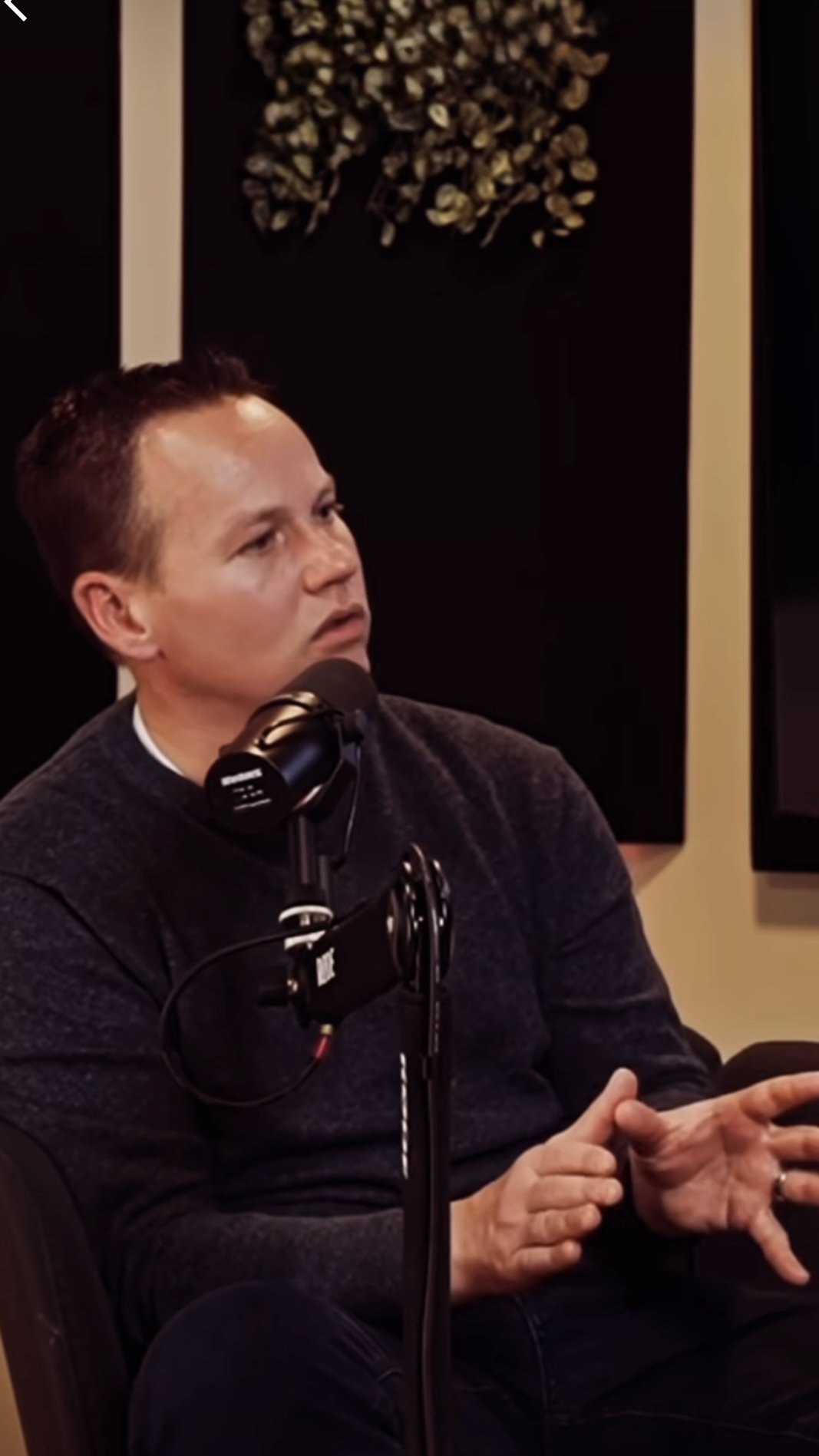 A man with short dark hair and light skin, wearing a dark sweater, sitting in front of a microphone in a recording studio or podcast setup, with soundproof walls and artwork behind him.