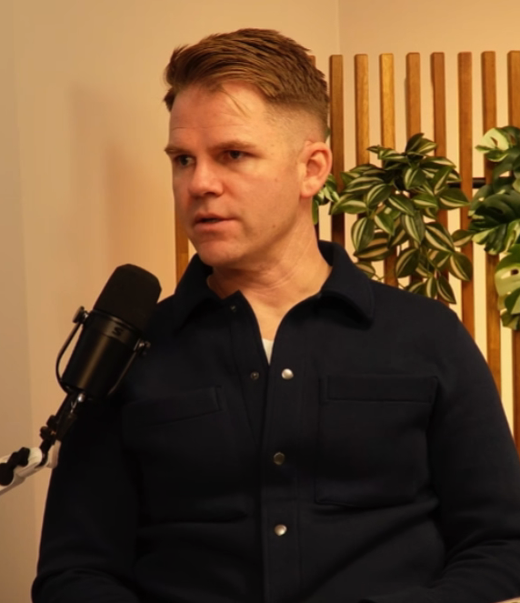 A man with short, light brown hair wearing a dark button-up shirt sitting in front of a microphone with a plant and wooden slats in the background.