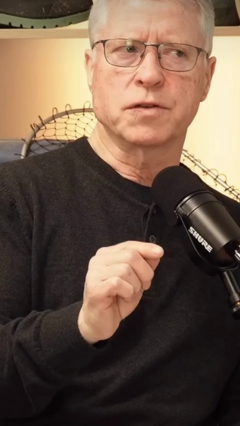 A man with glasses holding a microphone in front of a wall with a decorative hockey net and artwork.