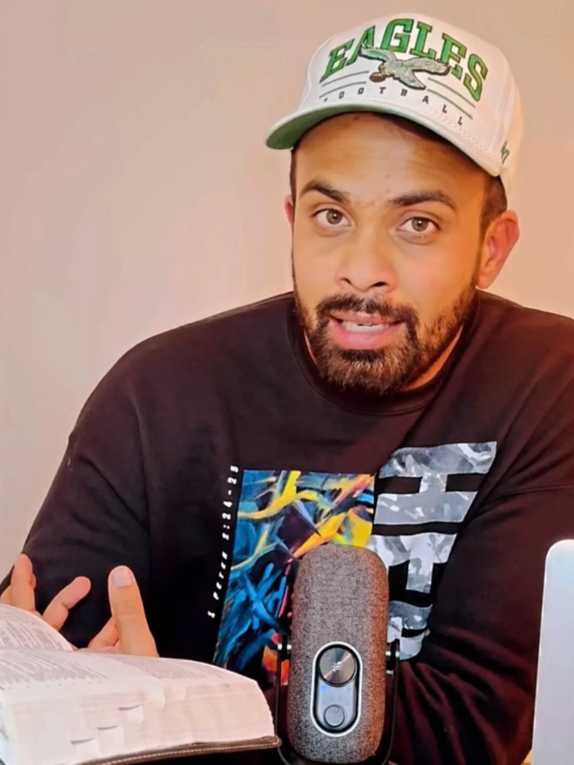 A man with a beard and short hair wearing a dark graphic T-shirt and a white Philadelphia Eagles baseball cap sitting at a table with a microphone, an open book, and a tablet in front of him, speaking directly to the camera.