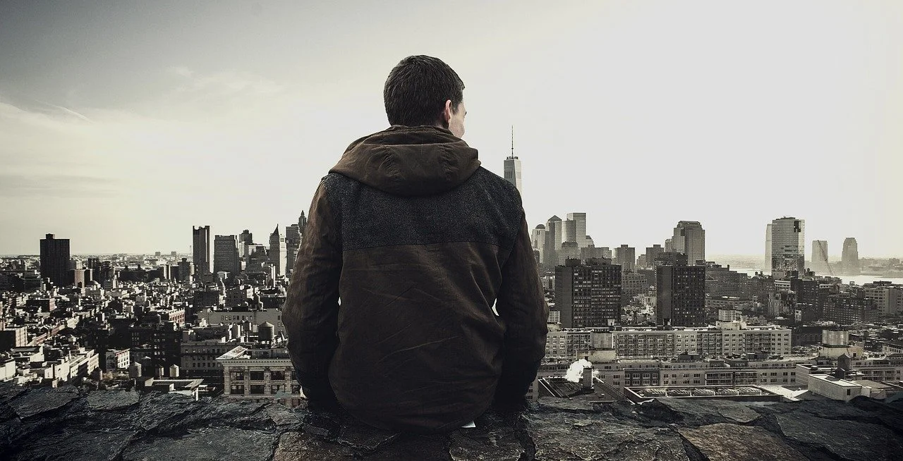A person sitting on a rooftop overlooking a city skyline with tall buildings and One World Trade Center in the distance.