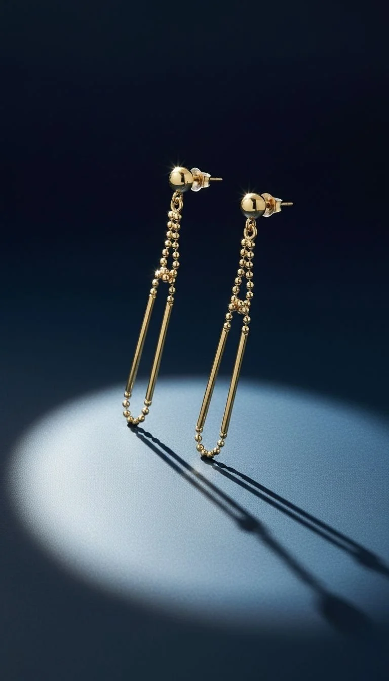 Gold dangling earrings with small beads and elongated bars, lit with a spotlight creating a shadow on a dark background.