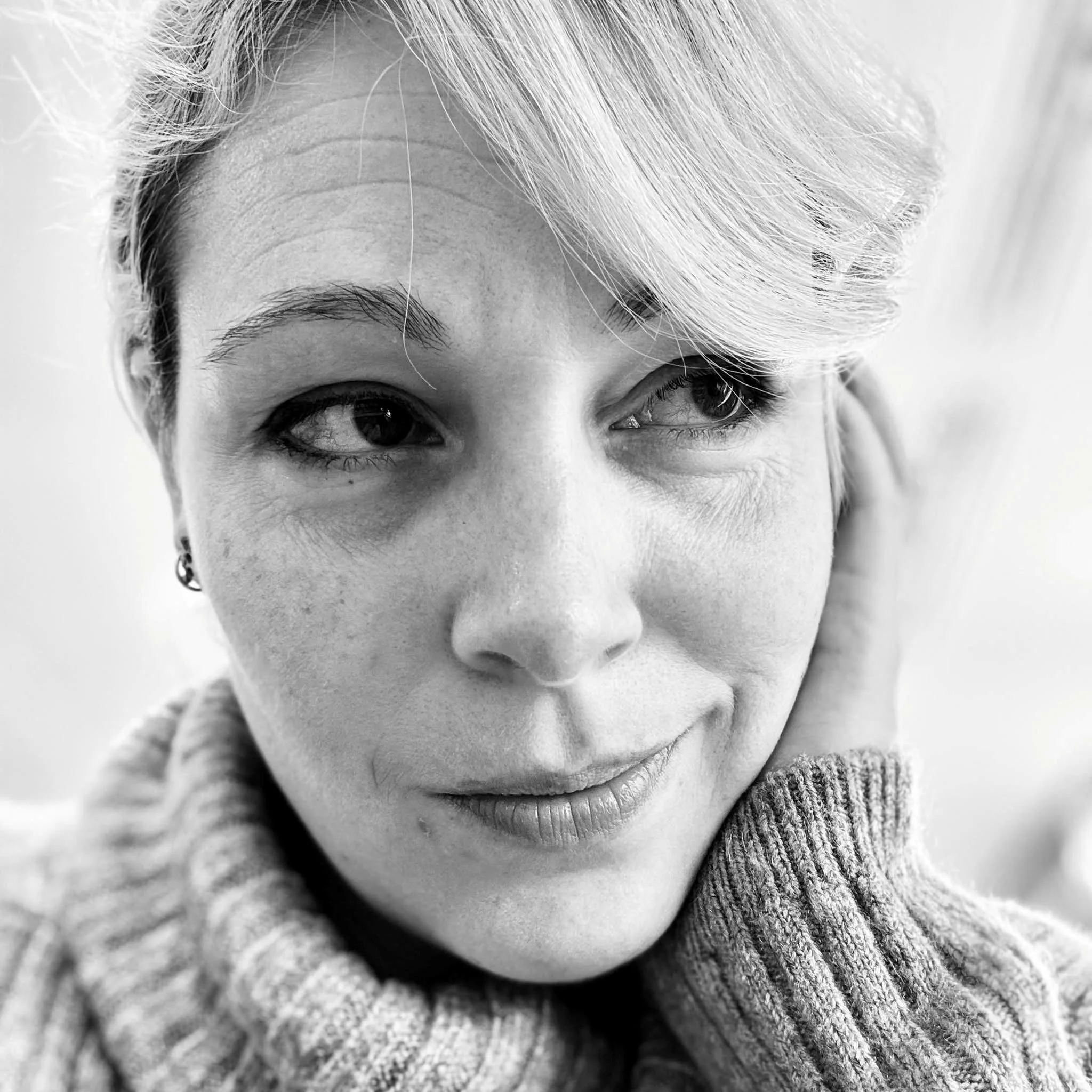 Close-up black and white portrait of a woman with short hair, wearing a knitted sweater, smiling softly and looking slightly to the side.