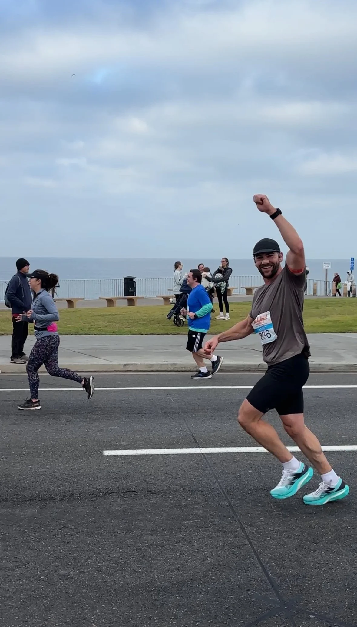 A man running in a marathon, smiling and raising his fist in celebration, with other runners and spectators along a seaside path under cloudy skies.