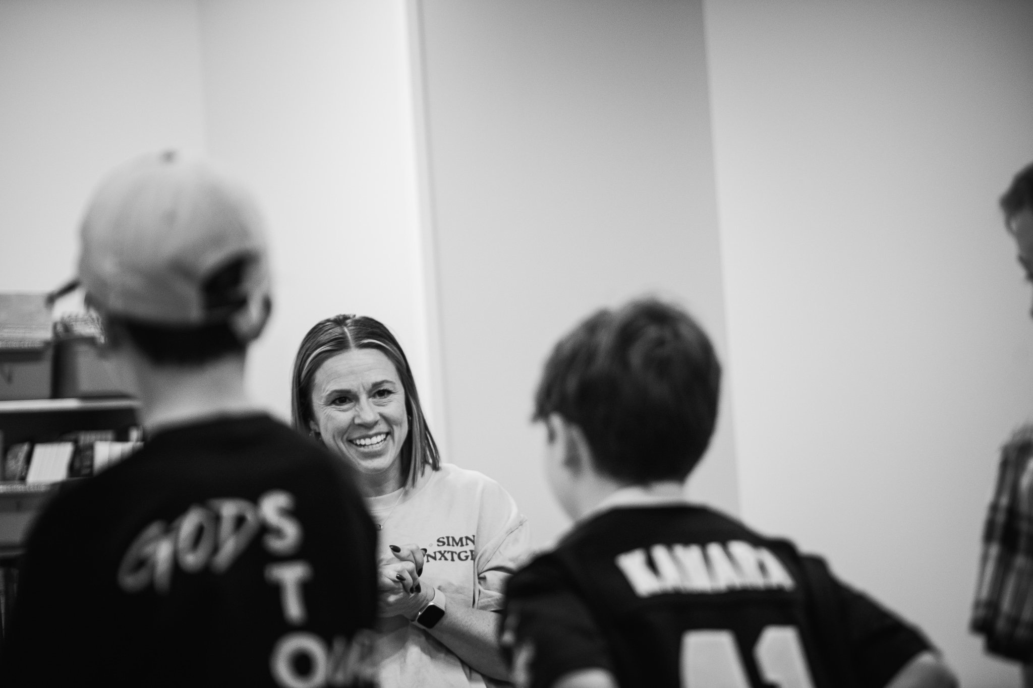 A man in a plaid shirt shows crayons in a plastic bin to a young boy, while another man with a name tag sits nearby. They are in a room with a table and black chairs, with blue walls and colorful equipment in the background.