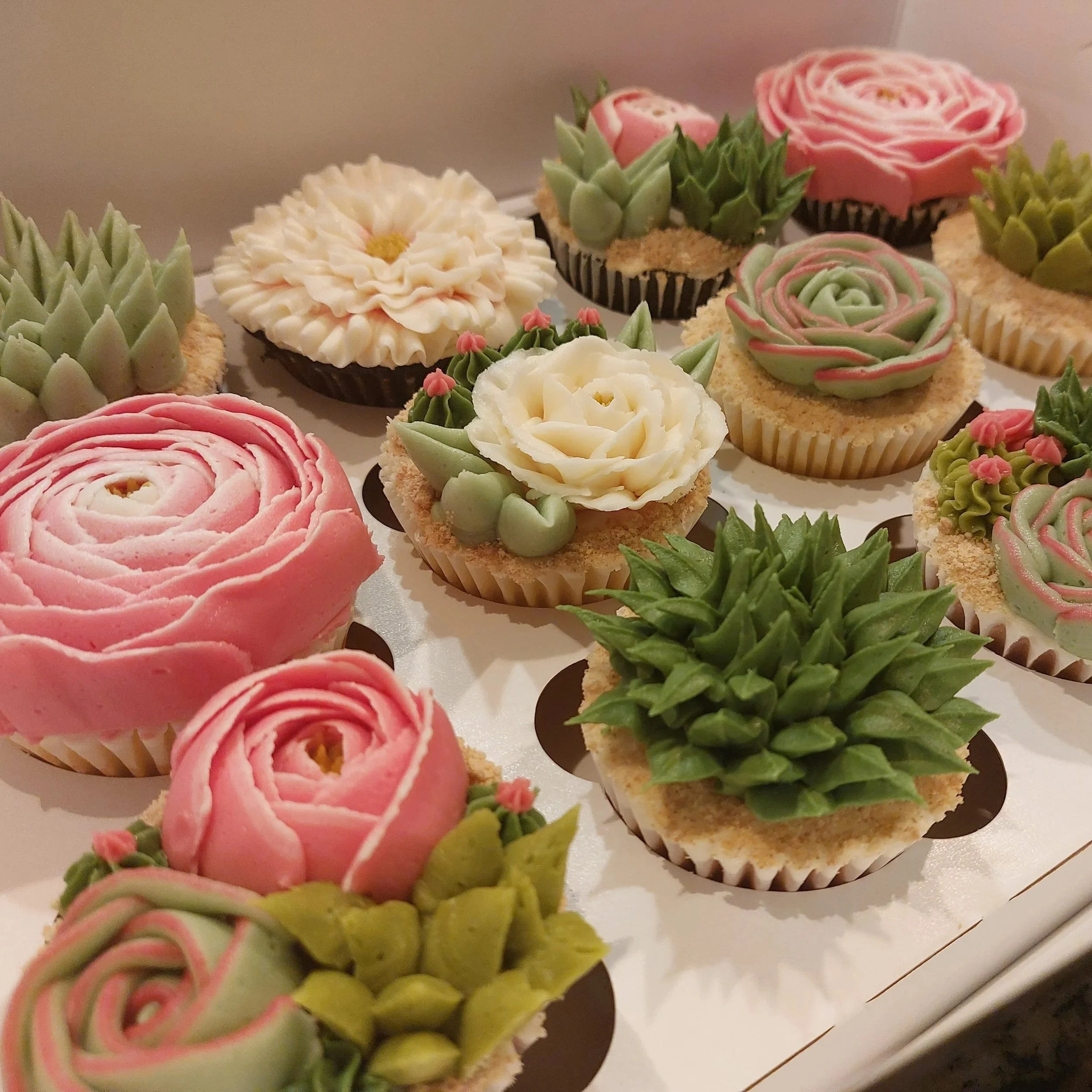 Decorative cupcakes with floral and succulent designs, in pastel pink, white, green, and beige colors.