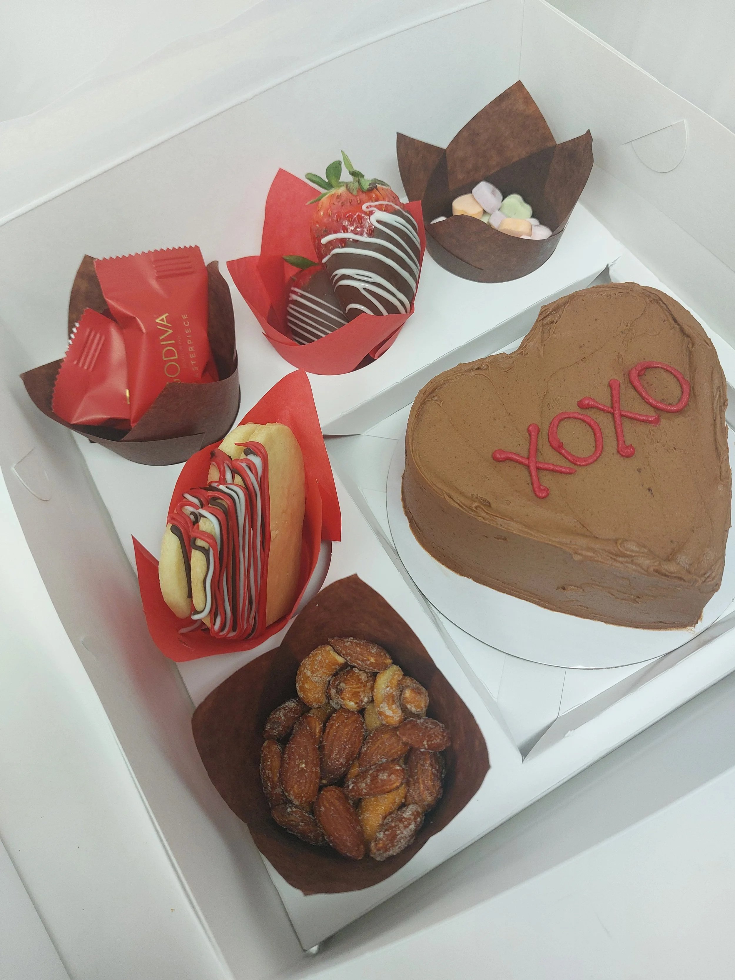 A box of assorted treats including a heart-shaped frosted cake with the word 'XOXO' on top, strawberries dipped in chocolate, a macaron topped with chocolate drizzle, a scoop of mixed candies, a chocolate-covered almond, and a piece of candy wrapped in red foil.