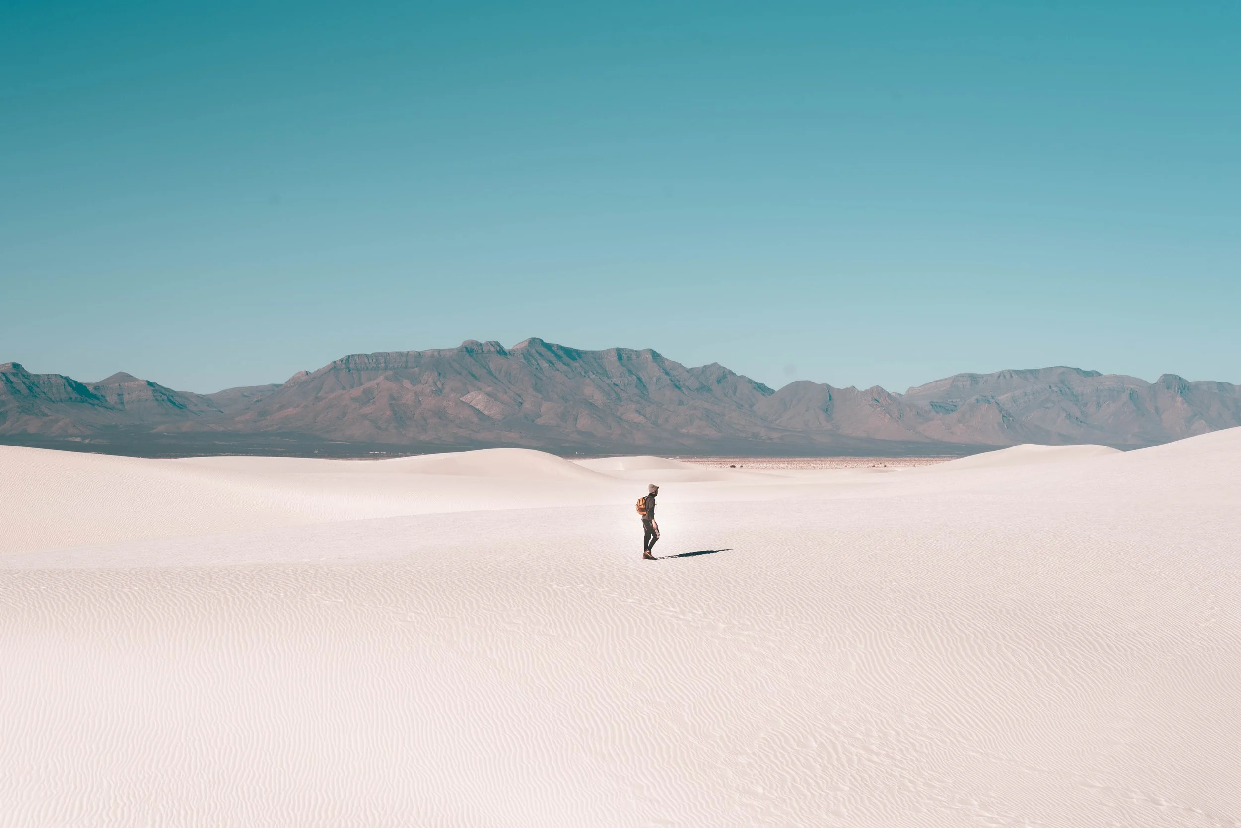 A person walking in a vast desert with white sand dunes and mountains in the background under a clear blue sky.