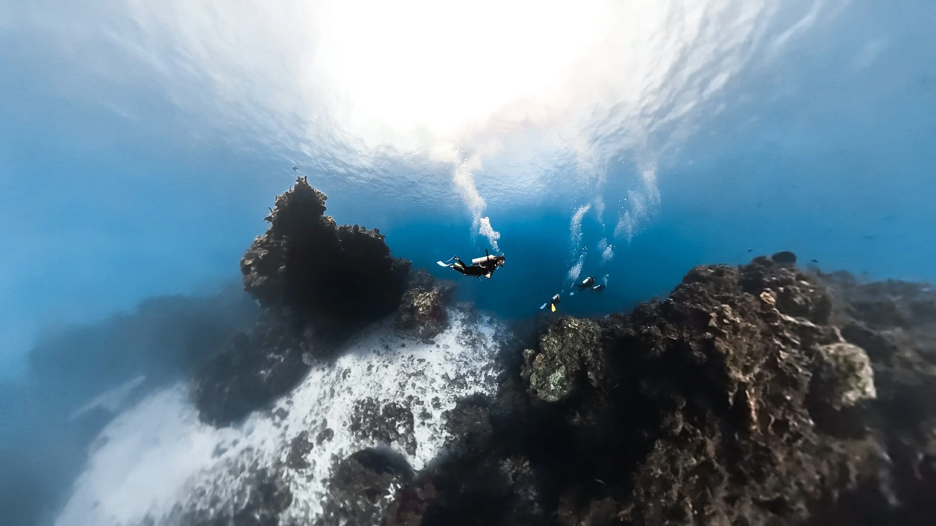 Underwater scene with a scuba diver swimming near rocks, sunlight filtering through the water, and bubbles rising toward the surface.