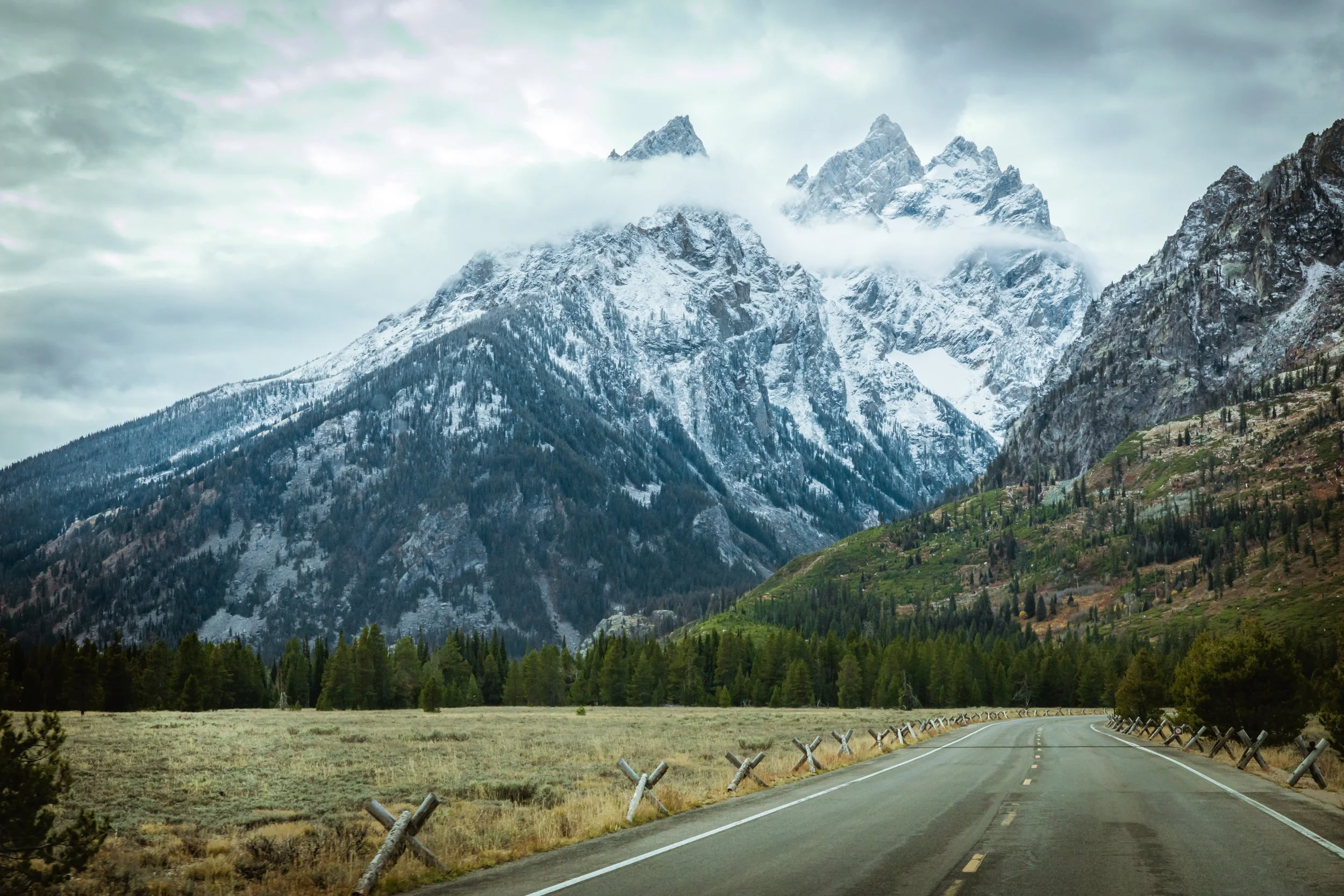 A winding road leading toward snow-capped mountains partially obscured by clouds, with green trees and grassy fields on the roadside.