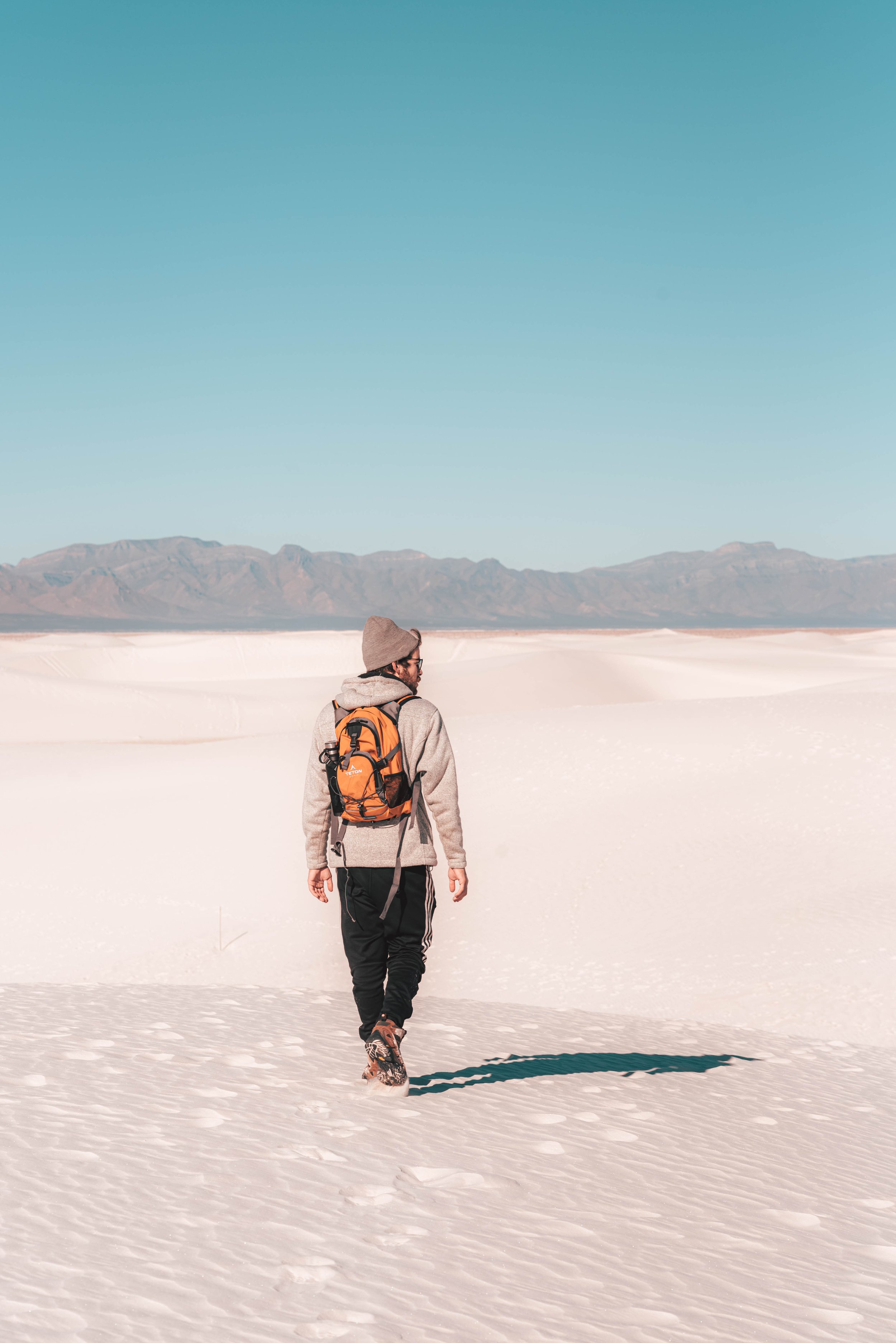 A man wearing a hoodie, beanie, sunglasses, and hiking boots walking through a snowy desert landscape with mountains in the background under a clear blue sky.