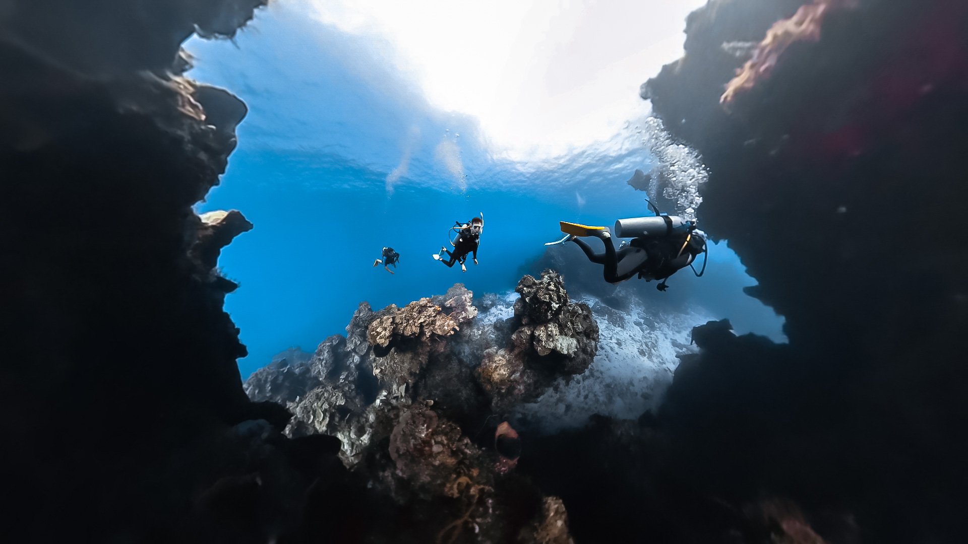 Underwater scene with three scuba divers exploring a rocky coral reef, with sunlight shining through the water from above.